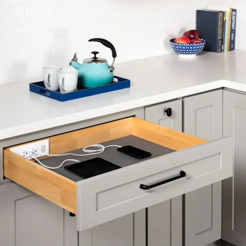 Open kitchen drawer with wooden interior, containing a power outlet, charging cable, black tablet, and smartphone, against a light gray kitchen cabinet.