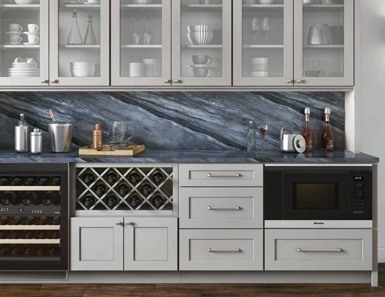 Modern kitchen with gray cabinets, glass-front upper cabinets, marble backsplash, wine rack, microwave, and various bottles on the countertop.