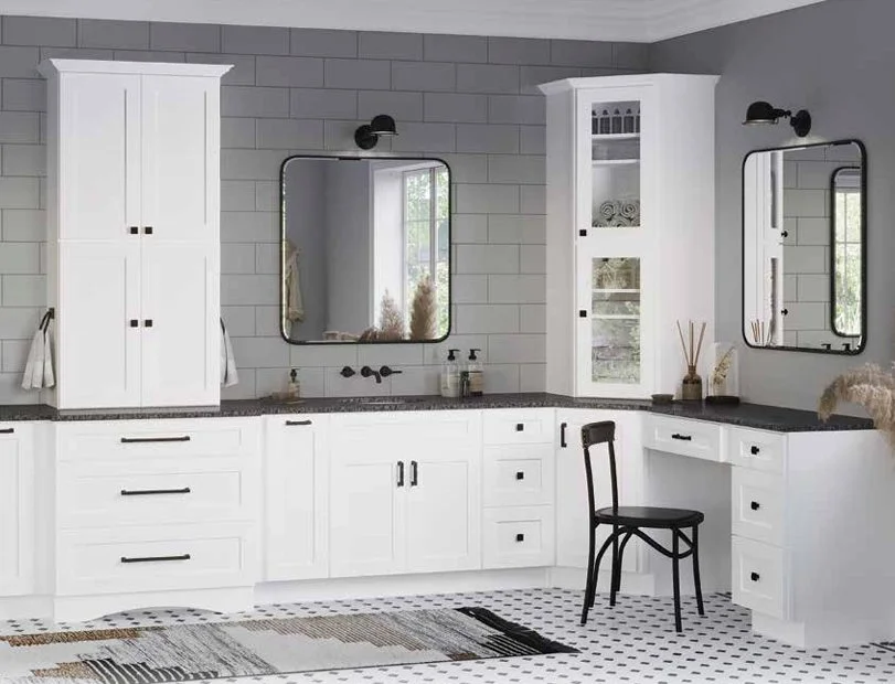 A modern bathroom with white cabinets and black hardware, two black framed mirrors above black countertops, and a black chair in front of a small white vanity. There are two windows, one between the mirrors and another in the corner, with decorative items and towels visible in the reflection and on the counters. The wall has neutral gray tiling and the floor features black and white patterned tiles.