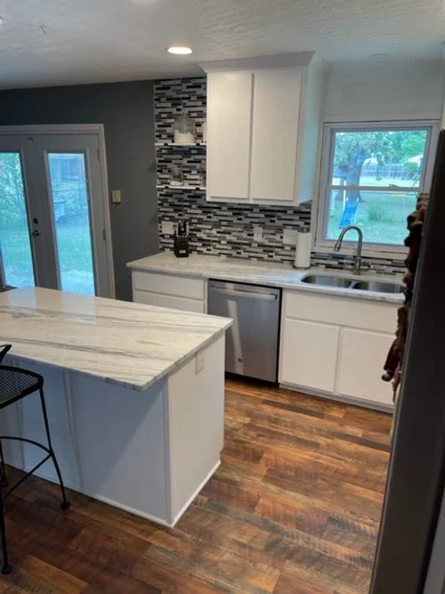 A modern kitchen with white cabinets, a marble countertop island, a window above the sink, and a decorative tile backsplash in shades of gray. There's a small fridge under the counter, hardwood floors, and a door leading outside.