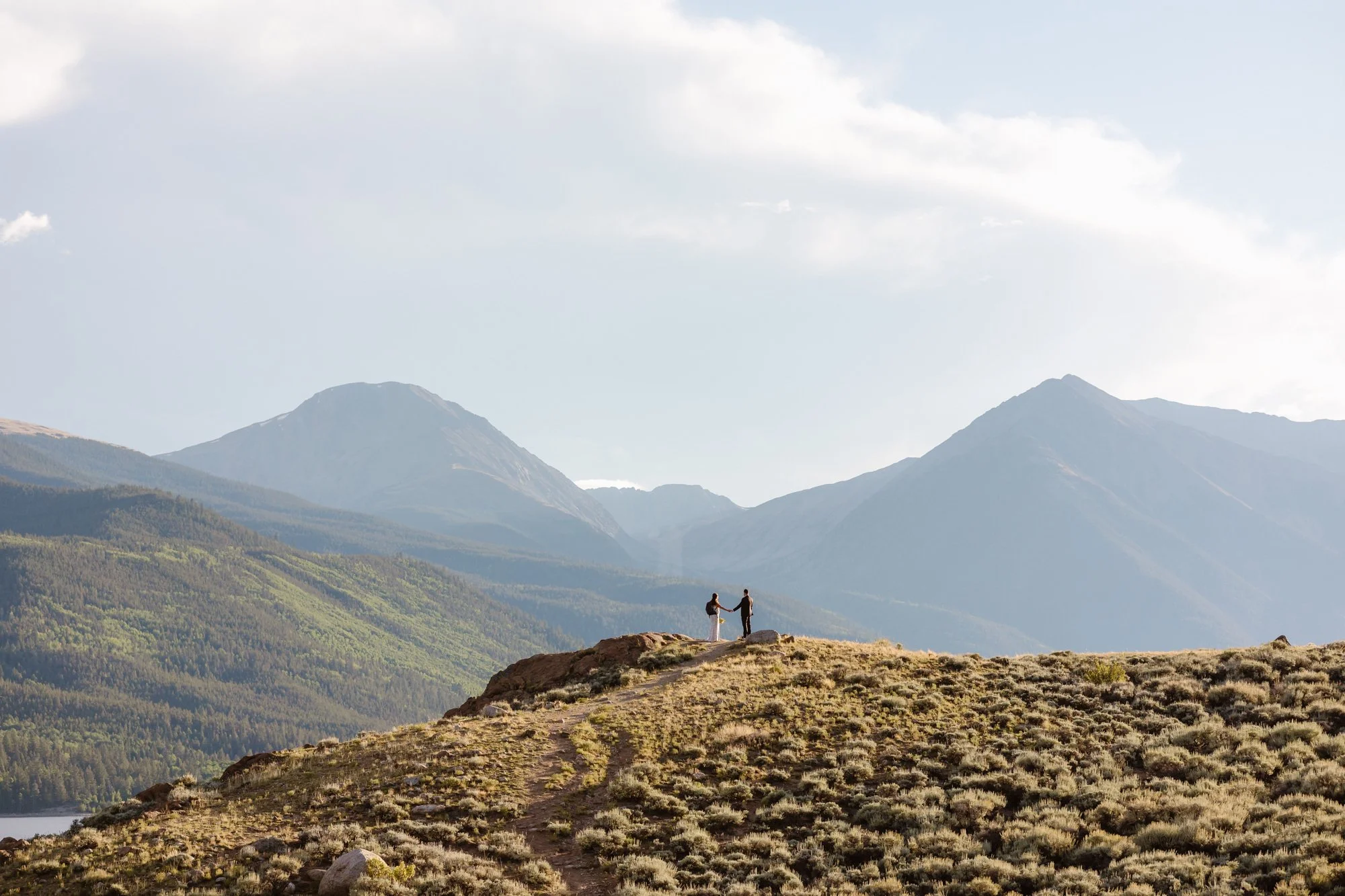 A couple holding hands on a mountain trail with mountain ranges and cloudy sky in the background.