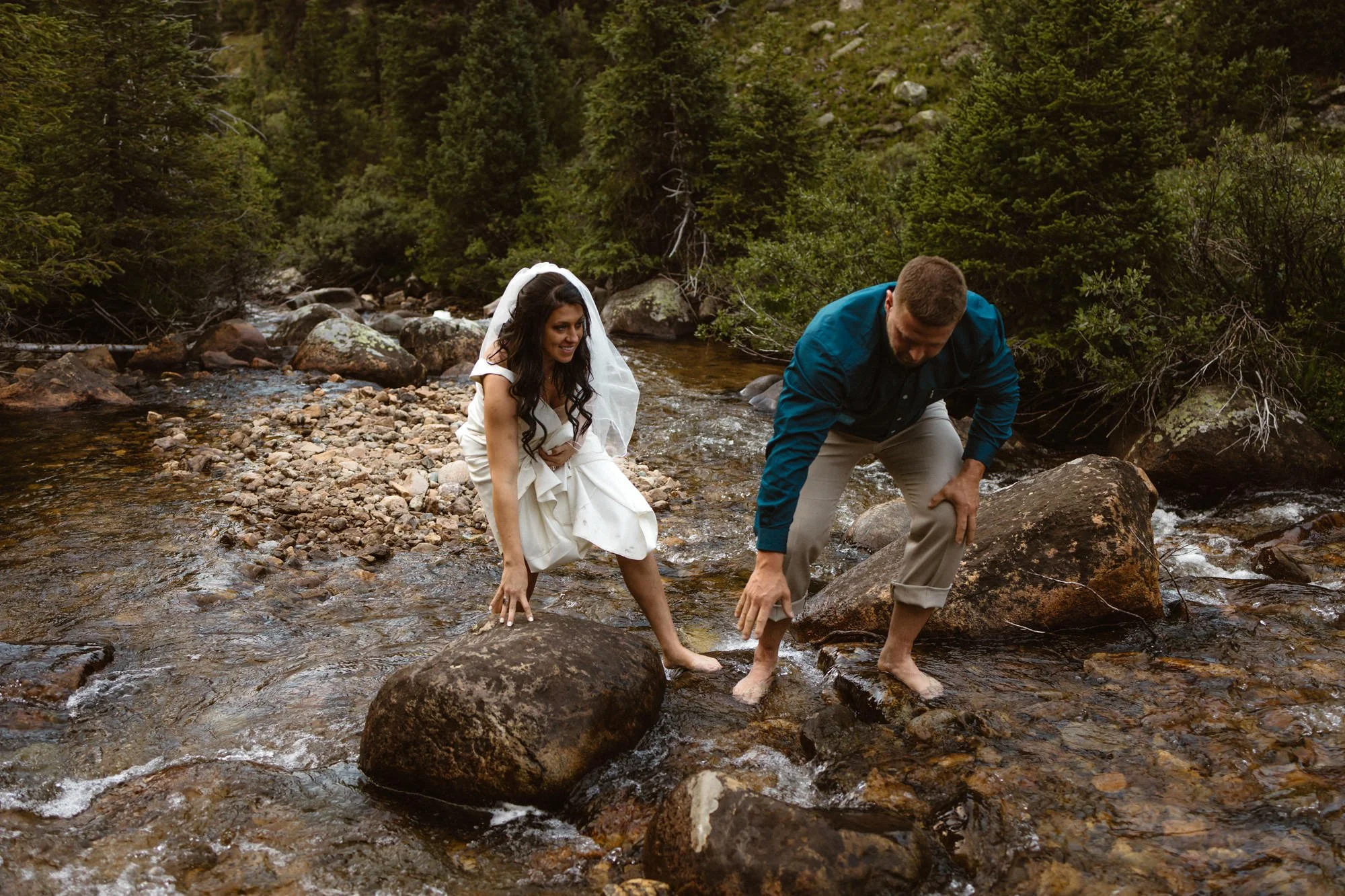 A woman in a wedding dress and a man in a blue shirt and beige pants wading through a shallow rocky stream in a forest.