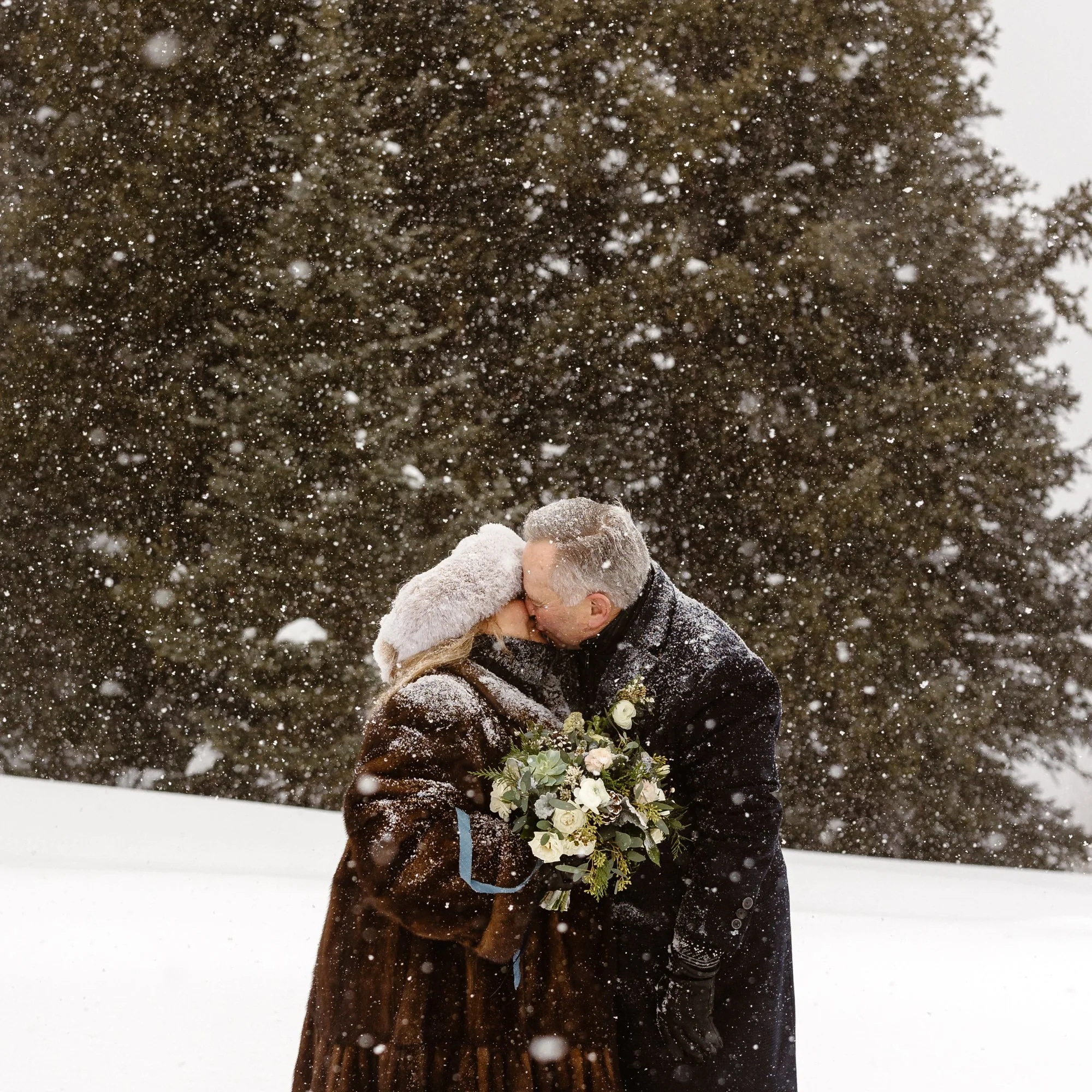 A couple kissing outdoors in the snow during a winter wedding, with the groom holding a bouquet of white flowers and greenery.