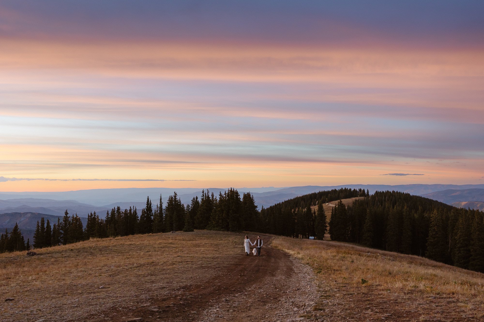 A couple holding hands while walking a dog on a dirt path through a mountainous landscape at sunset, with colorful elongated clouds in the sky and evergreen trees in the background.