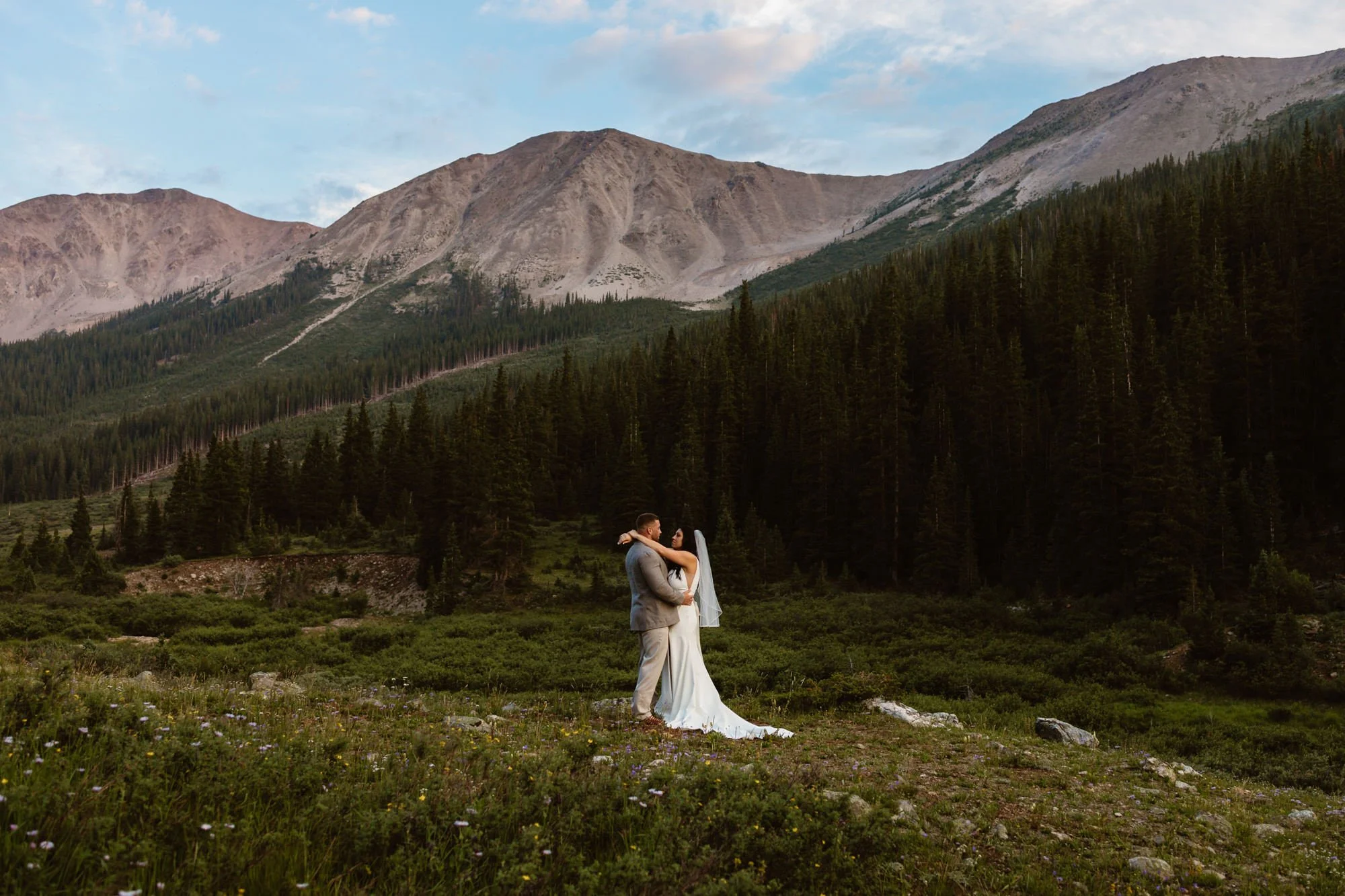 A bride and groom embracing outdoors in a mountainous area with a large mountain range and evergreen forest in the background, and a partly cloudy sky overhead.