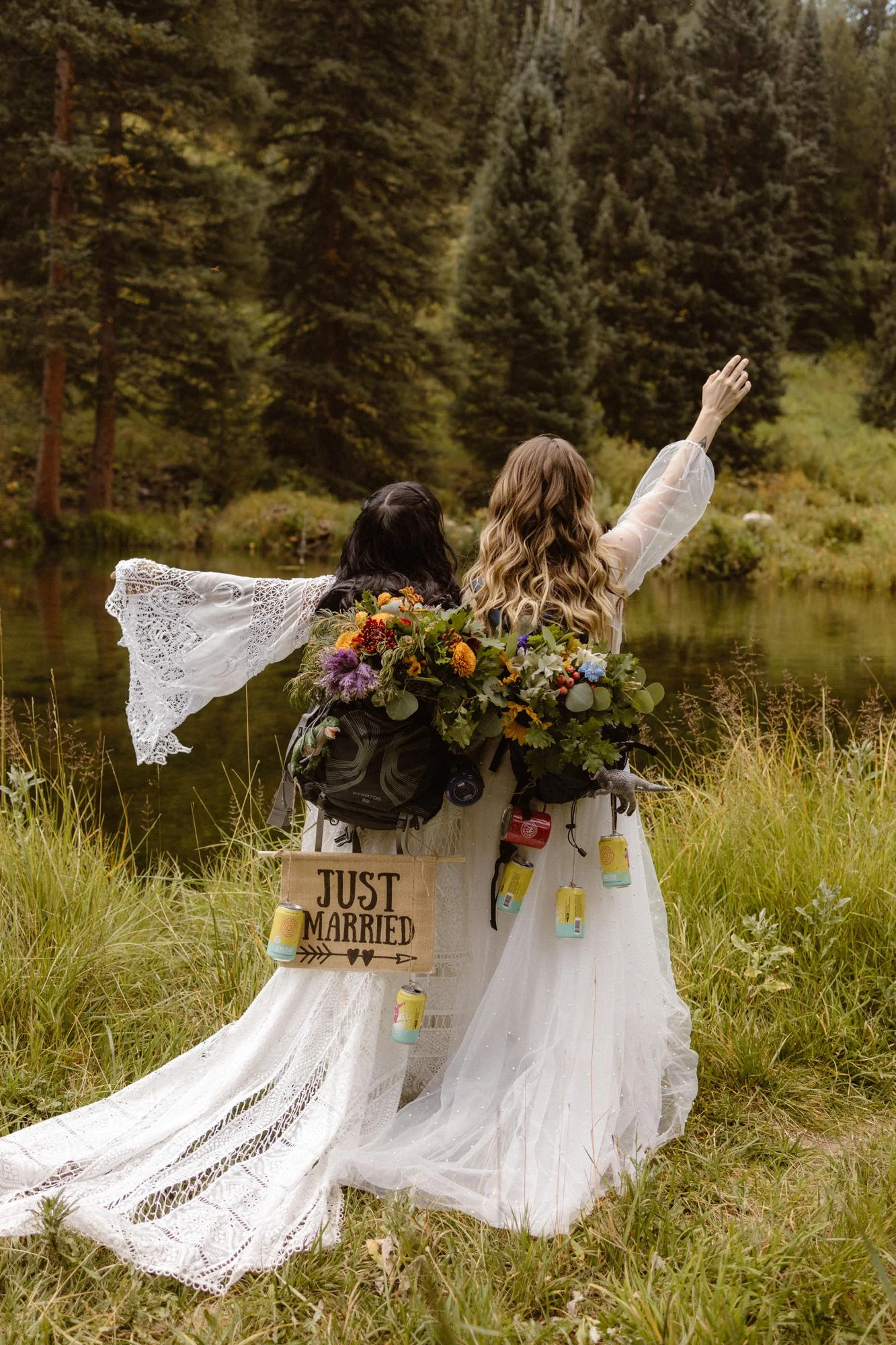 Two women in wedding dresses on a grassy riverbank, with one raising her hand, surrounded by trees and nature, and decorated with a sign that reads 'Just Married' and hanging drinks.