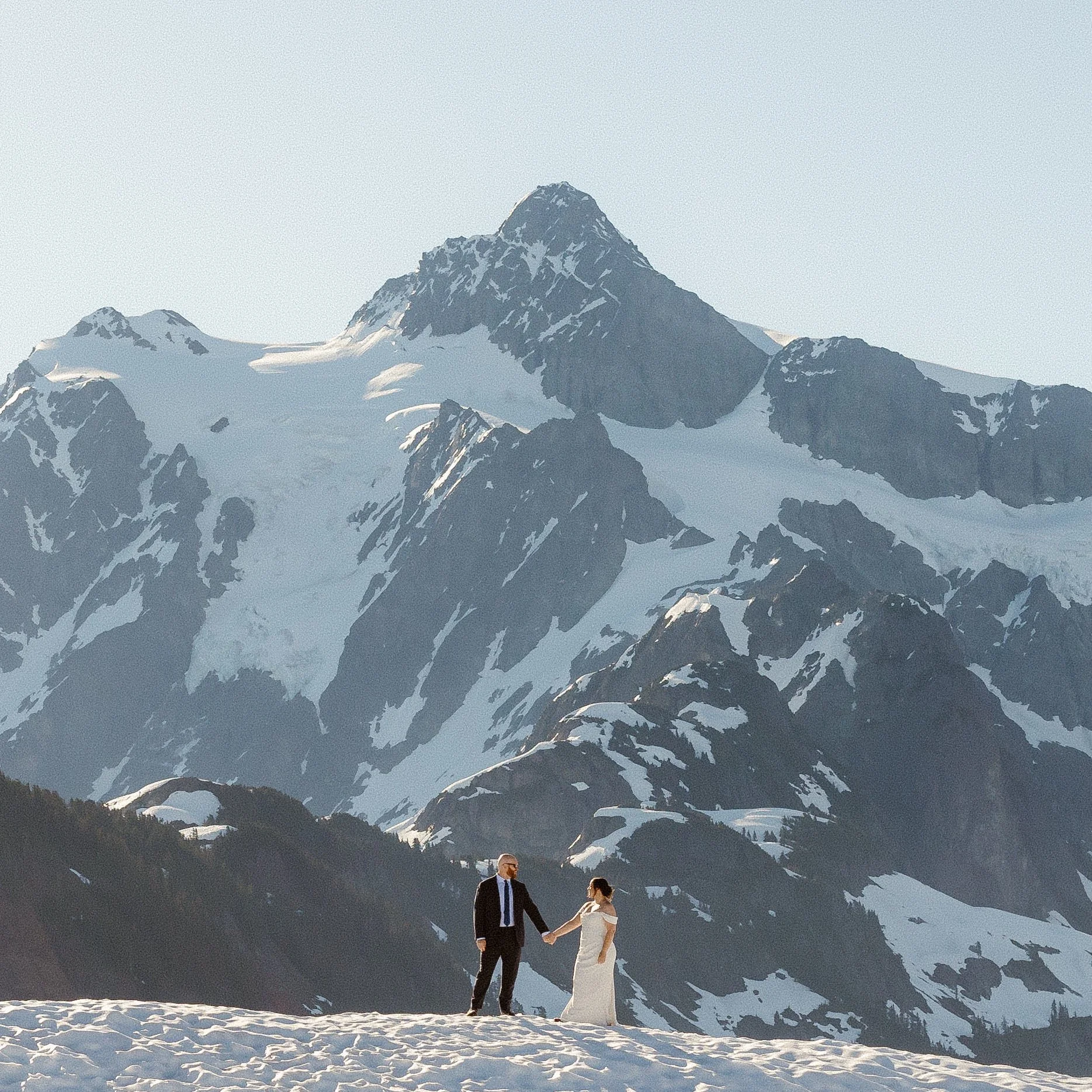 A man and woman holding hands in a snowy landscape with a mountain range in the background. The man is wearing a suit and sunglasses, and the woman is in a white dress. Snow covers the ground and mountain peaks under a clear sky.