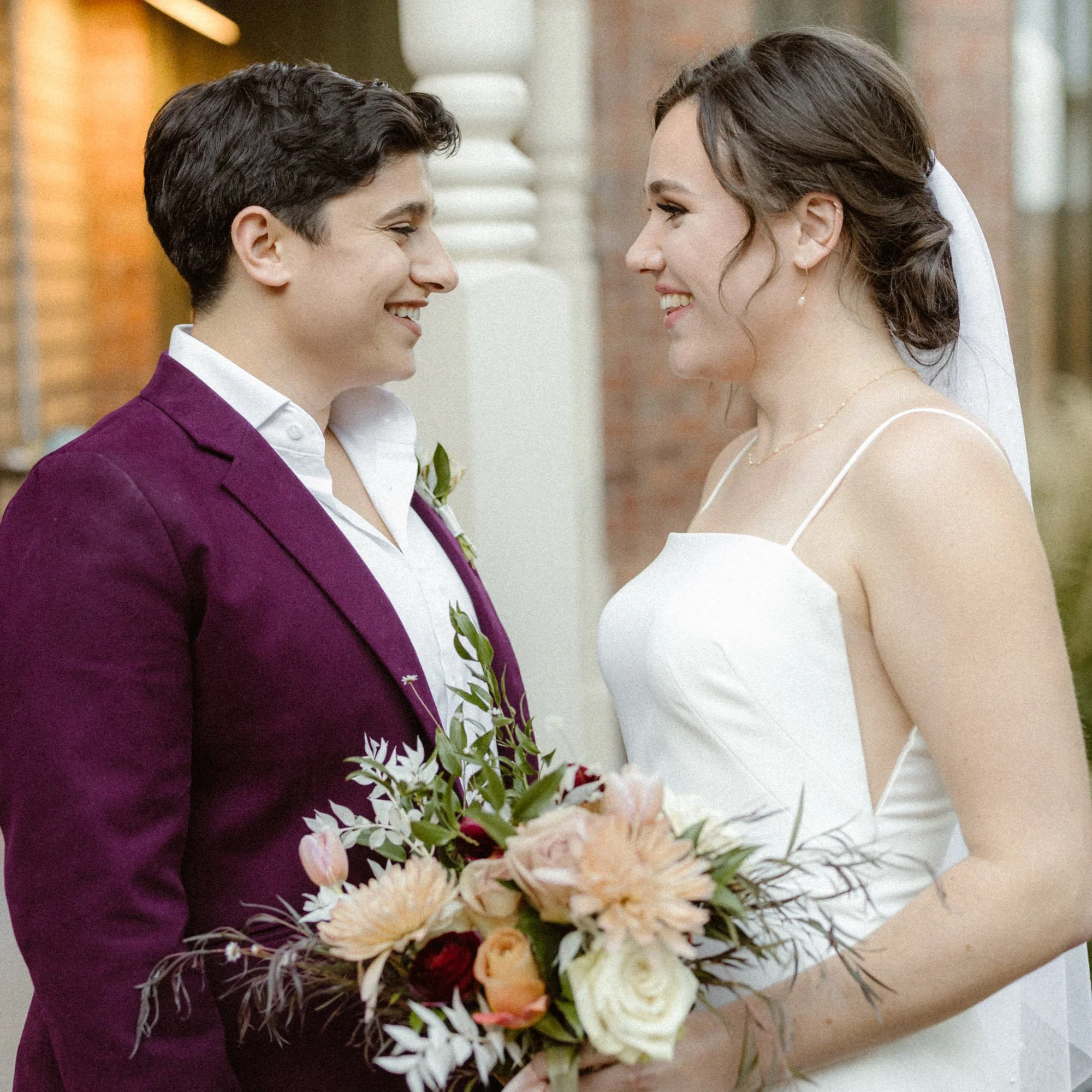 A couple at their wedding, standing face to face and smiling, holding a large bouquet of flowers.