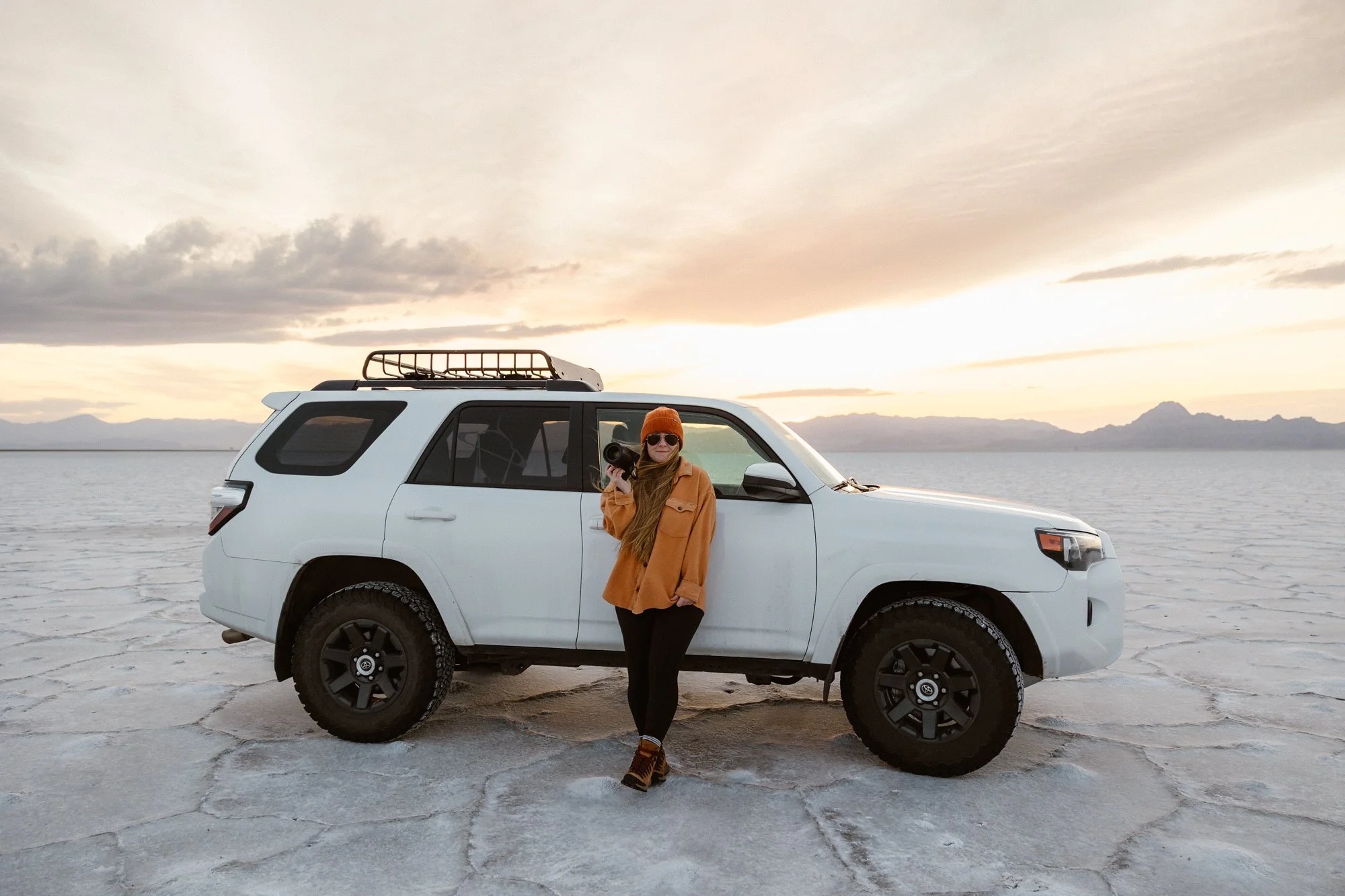 A woman wearing a brown jacket, orange beanie, sunglasses, and hiking boots stands next to a white SUV with a roof rack in a flat, salt-flat landscape during sunset or sunrise, with mountains in the distance and a cloudy sky.