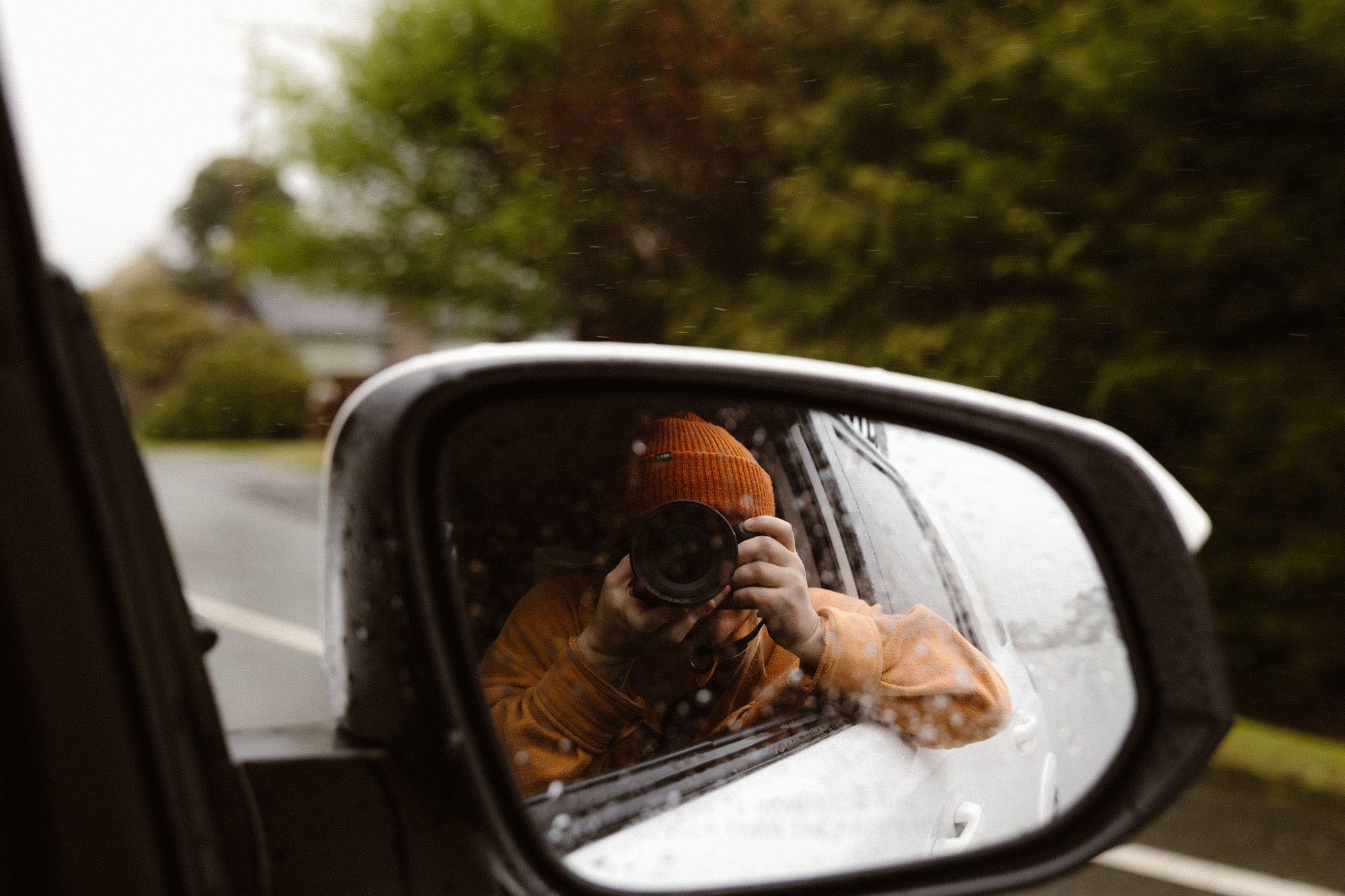 Person wearing an orange knit hat and orange jacket taking a photo with a camera in a car's side mirror on a rainy day.