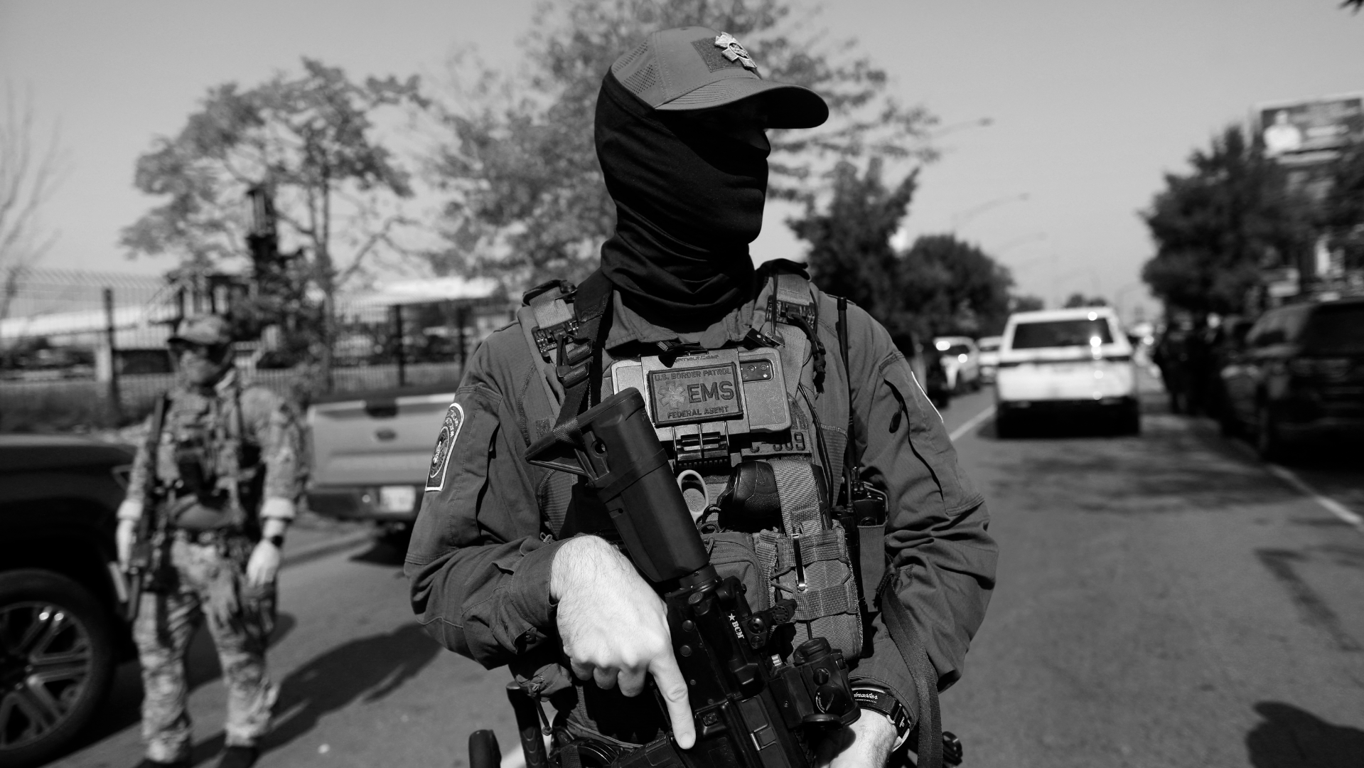 Black and white photo of a heavily armed officer in tactical gear, wearing a face covering and cap, standing on a street with cars and another officer in the background.
