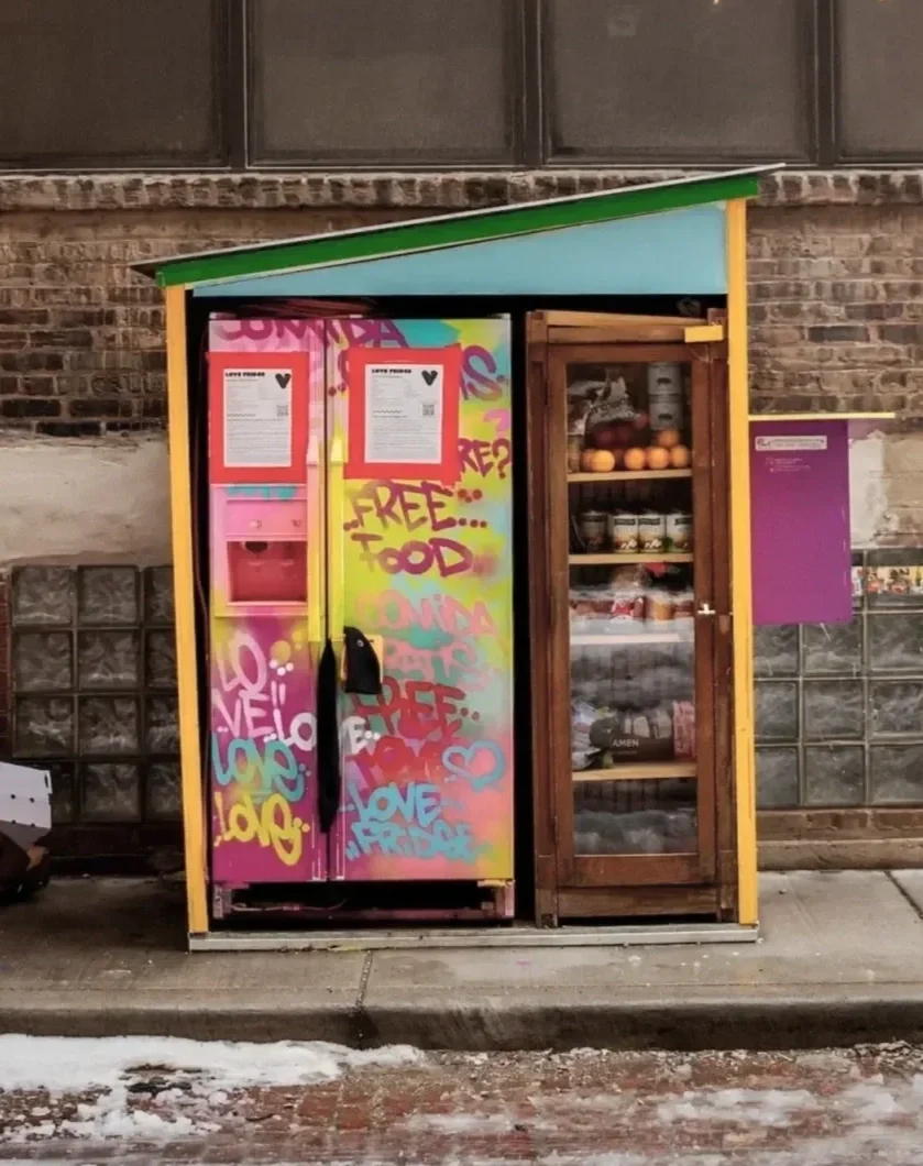 A small outdoor food stand with colorful graffiti on the door, displaying a sign for free food. Next to it, there is a glass cabinet filled with produce and other food items. The stand is set against a brick wall on a sidewalk with some snow and ice.