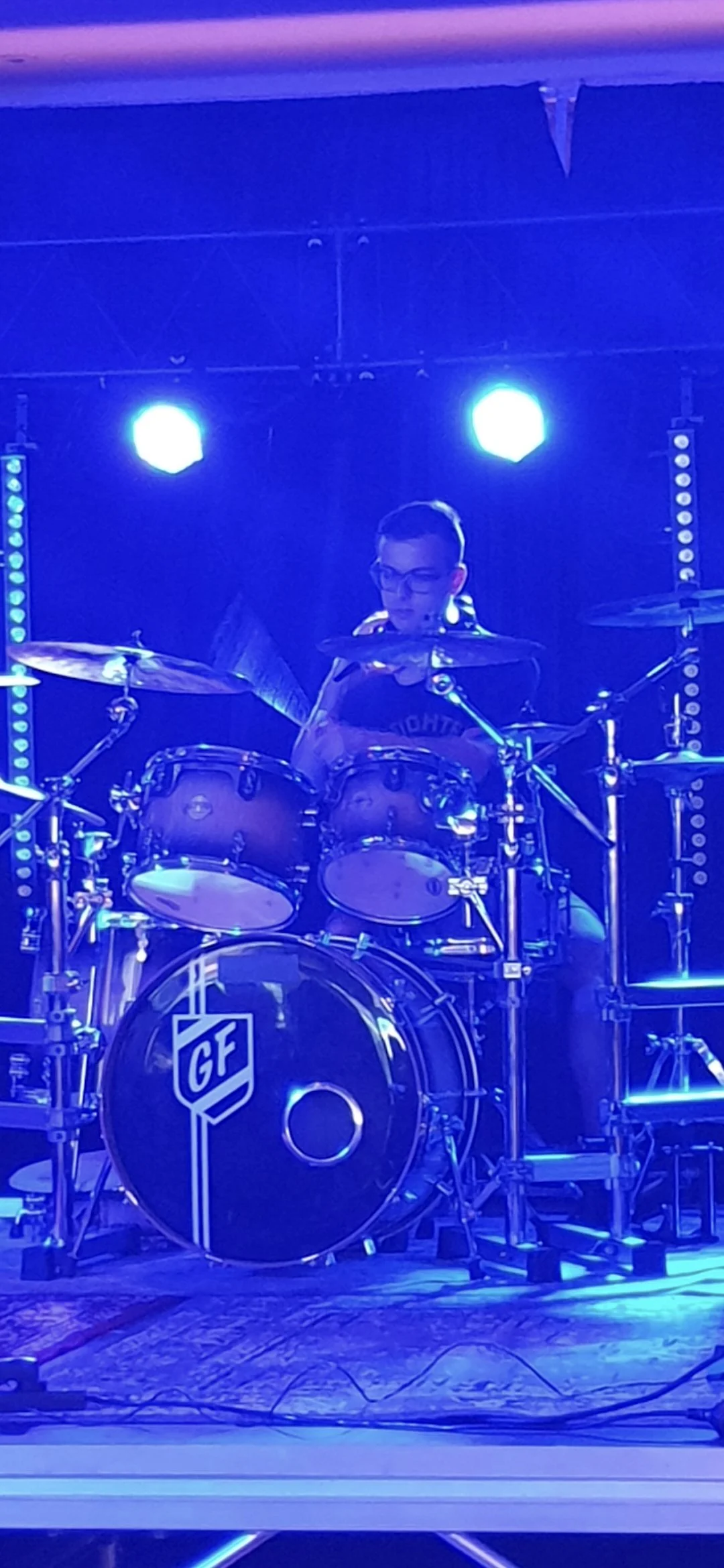 A person playing drums on stage with blue lighting and stage equipment in the background.