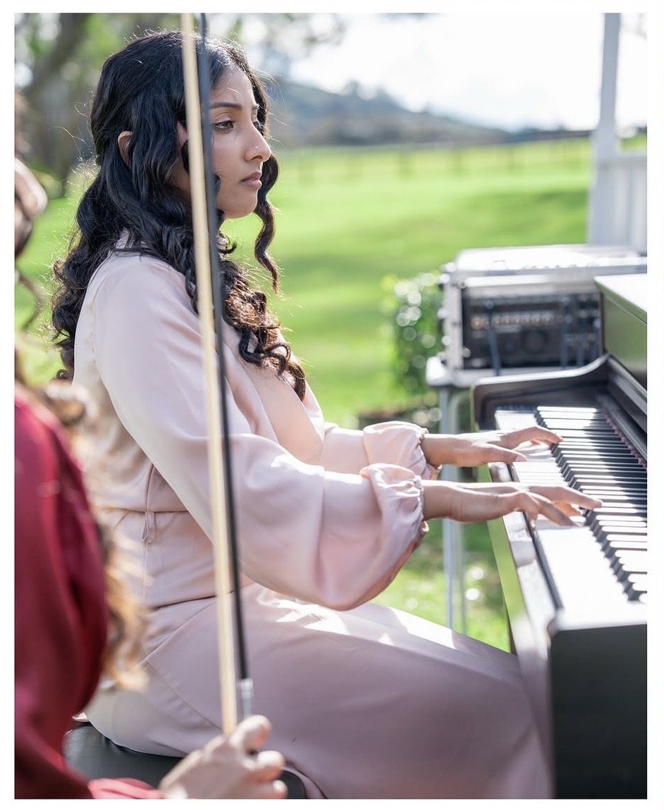 A woman playing the piano outdoors with a woman holding an umbrella nearby.