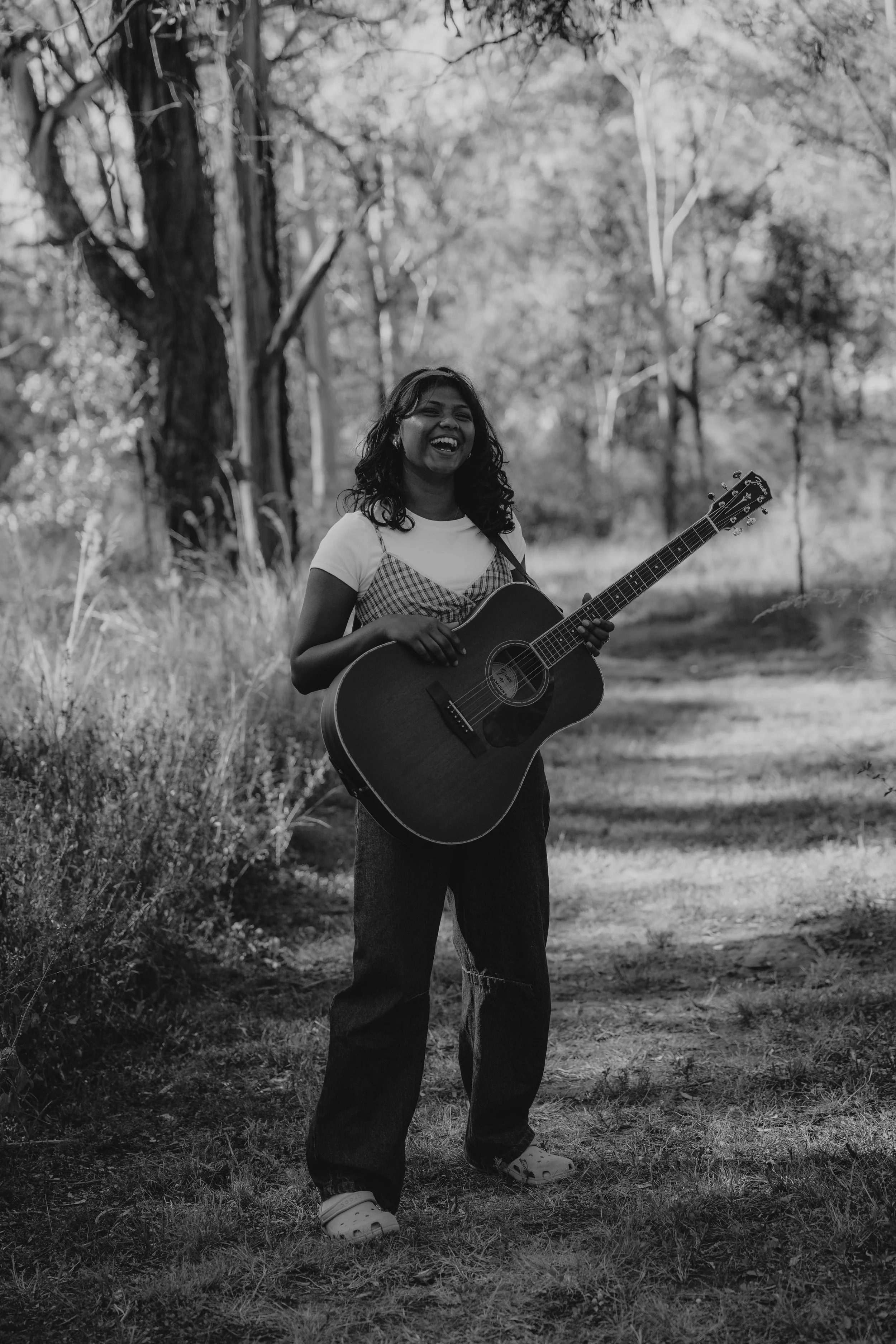 A woman with curly hair, smiling and singing while playing an acoustic guitar outdoors in a wooded area.