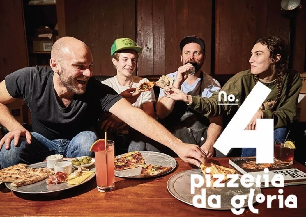 Group of four restaurant workers sitting at a table in a pizzeria, sharing pizza and drinks, with one person passing pizza to another. There are various pizzas, a cocktail, and other snacks on the table, in a cozy, wooden-paneled restaurant setting.