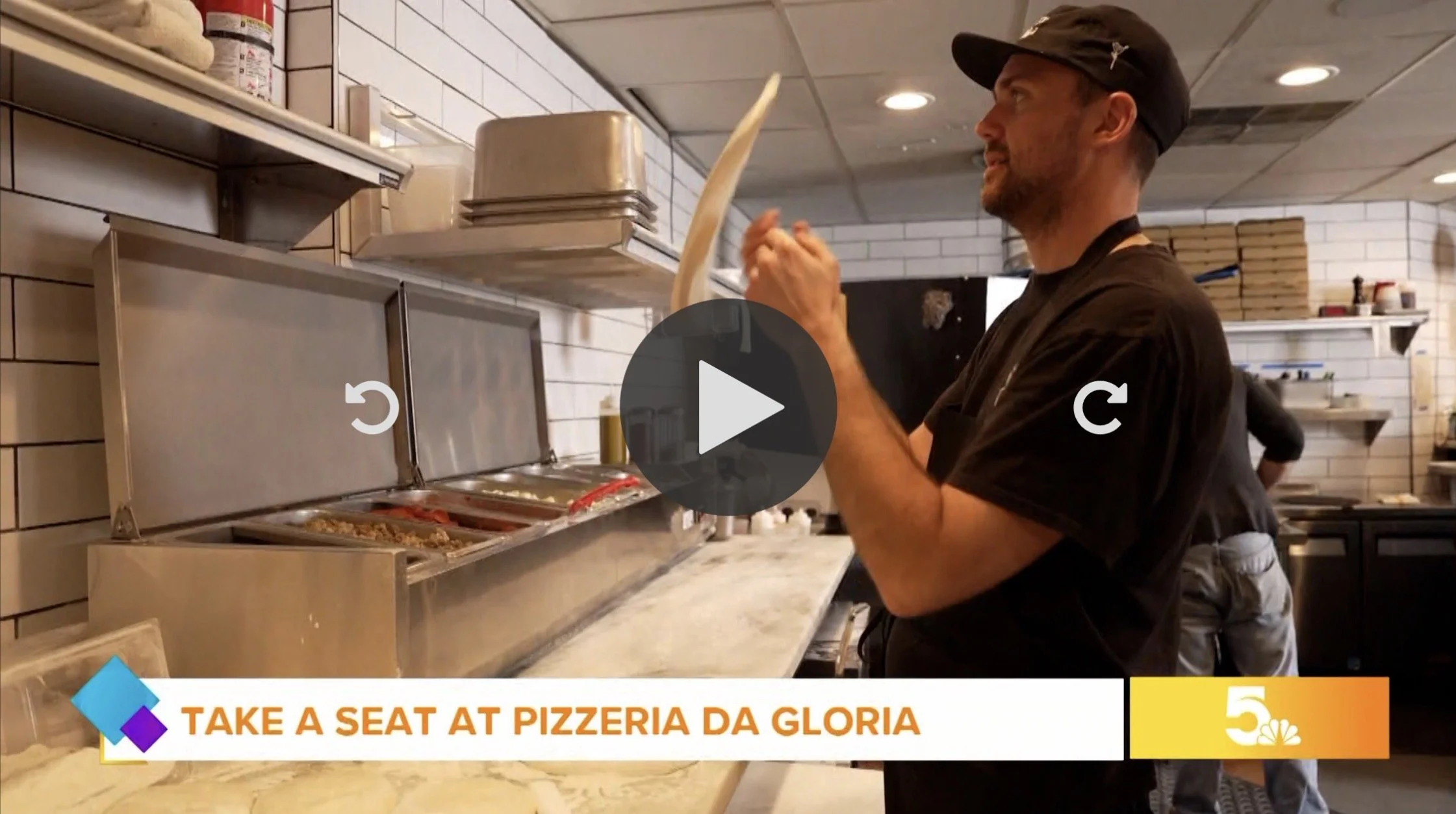 A man wearing a black cap and black shirt in a pizzeria kitchen, preparing dough or ingredients behind a silver counter with a food warmer and sauce containers. The kitchen has white tiled walls and shelves with pizza boxes, utensils, and supplies.