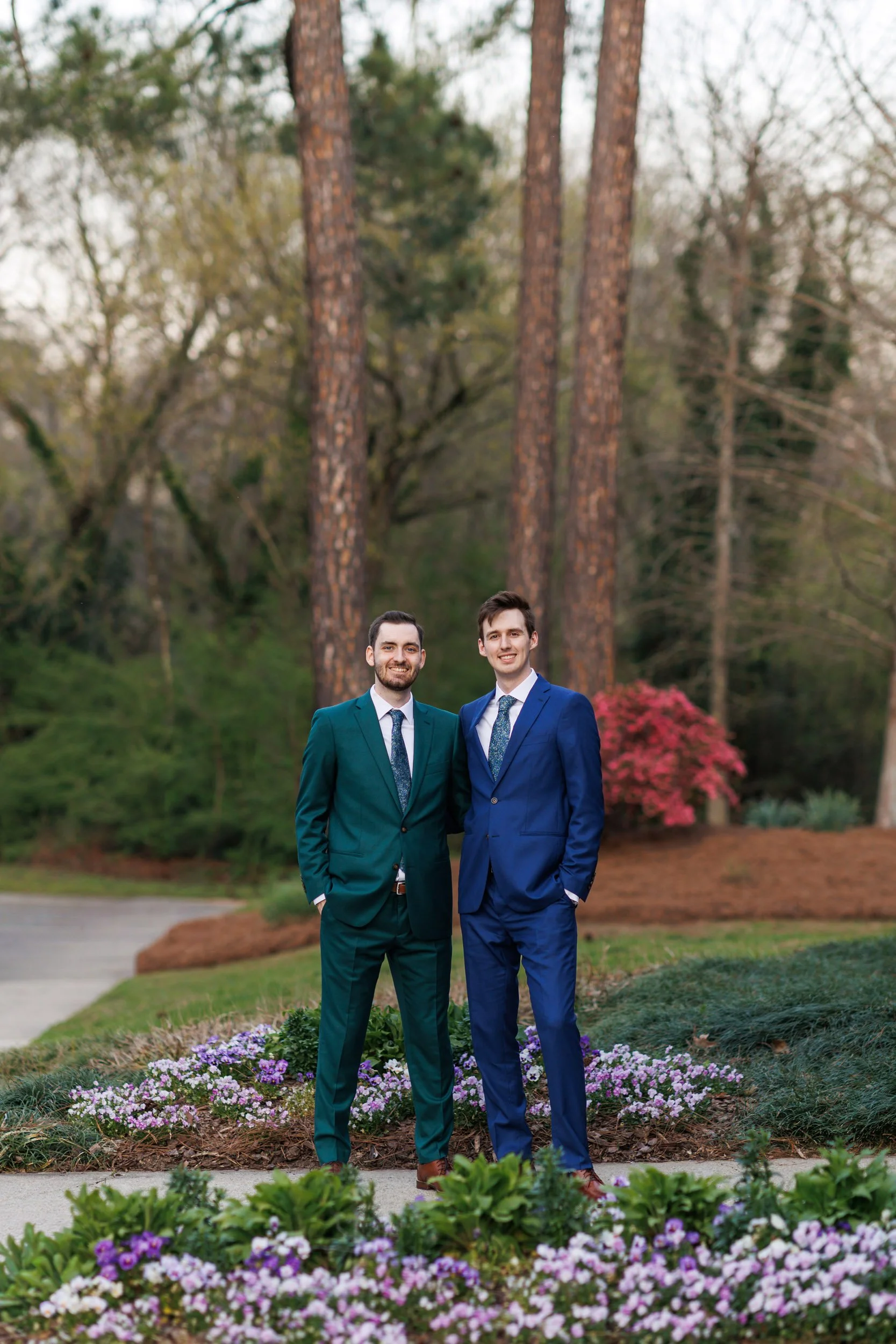 Two grooms in green and navy suits standing together in a garden at Roswell River Landing in Roswell, Georgia