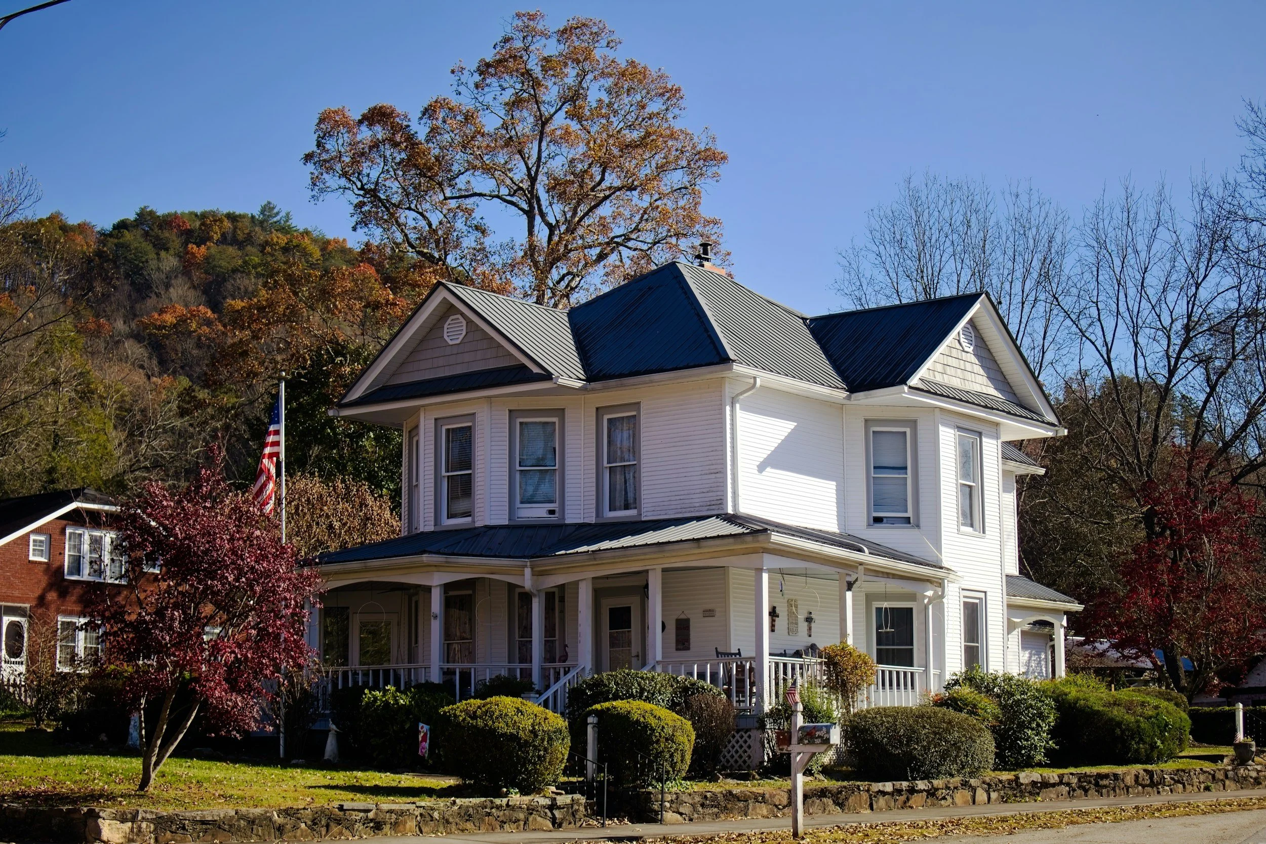 White two-story home with a wraparound porch and an American flag, surrounded by autumn trees under a blue sky