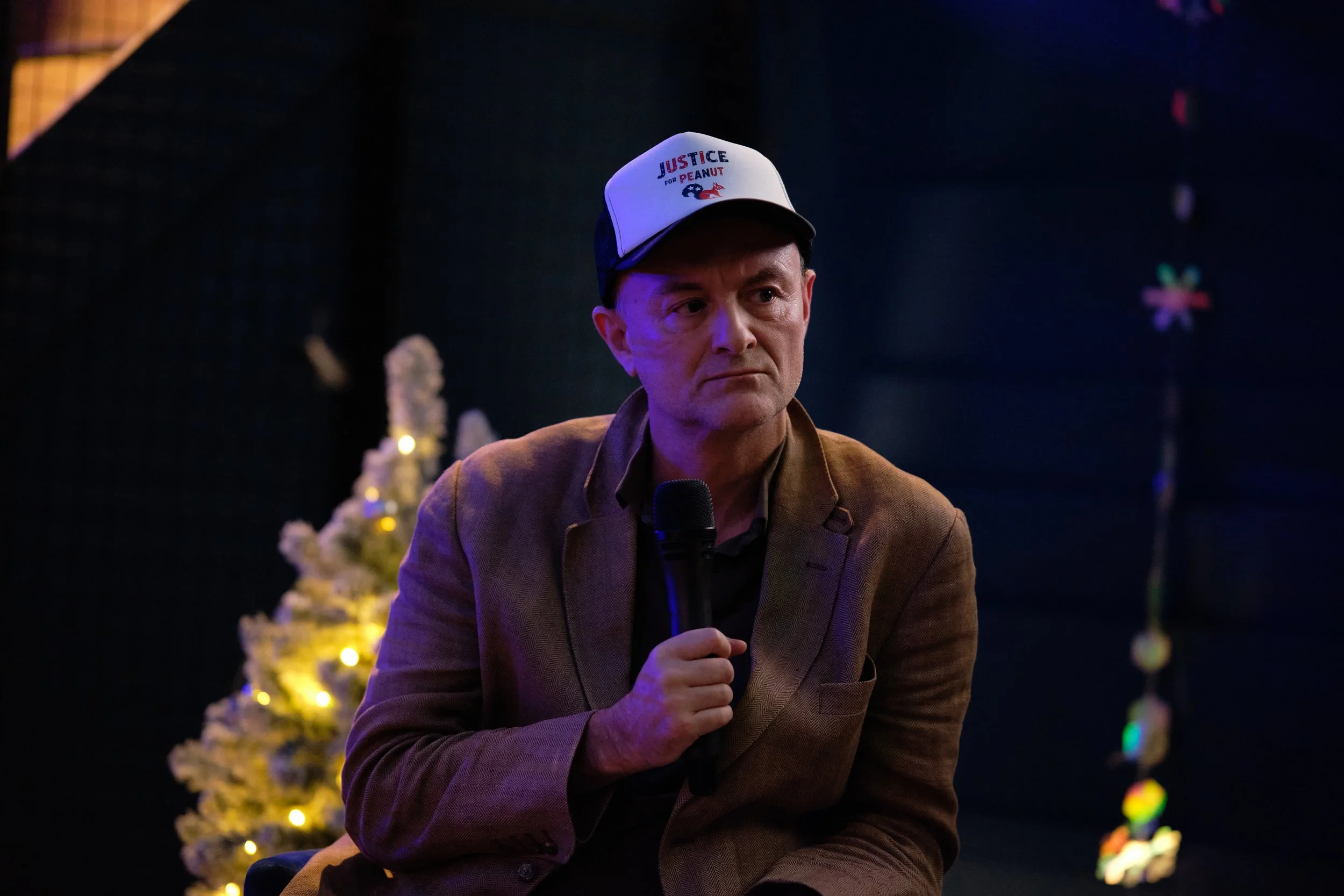 Man in a hat holding a microphone, sitting in front of a Christmas tree with lights.