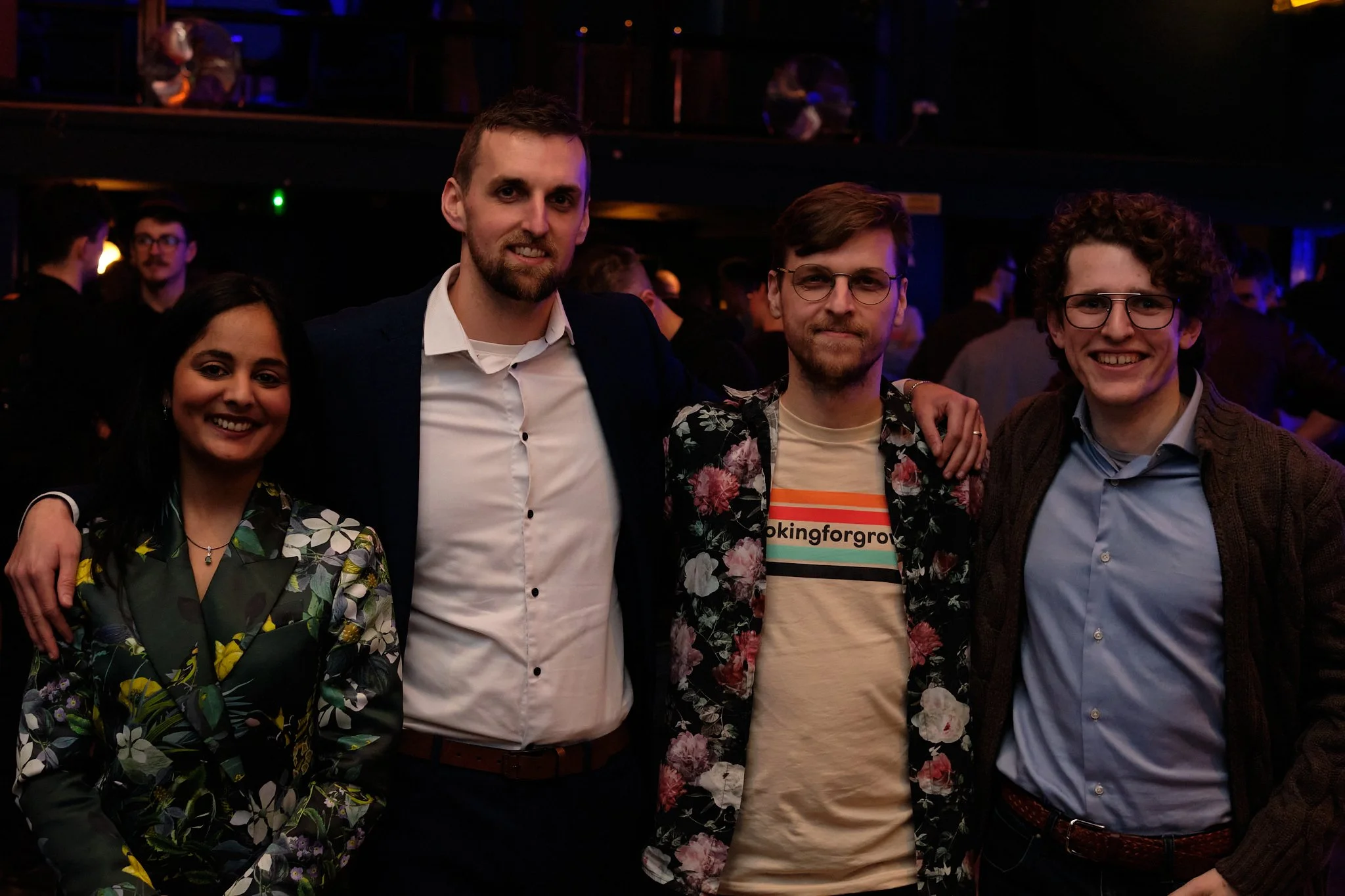 Group of four people posing together at an indoor event with a dimly lit background, wearing formal and semi-casual attire.
