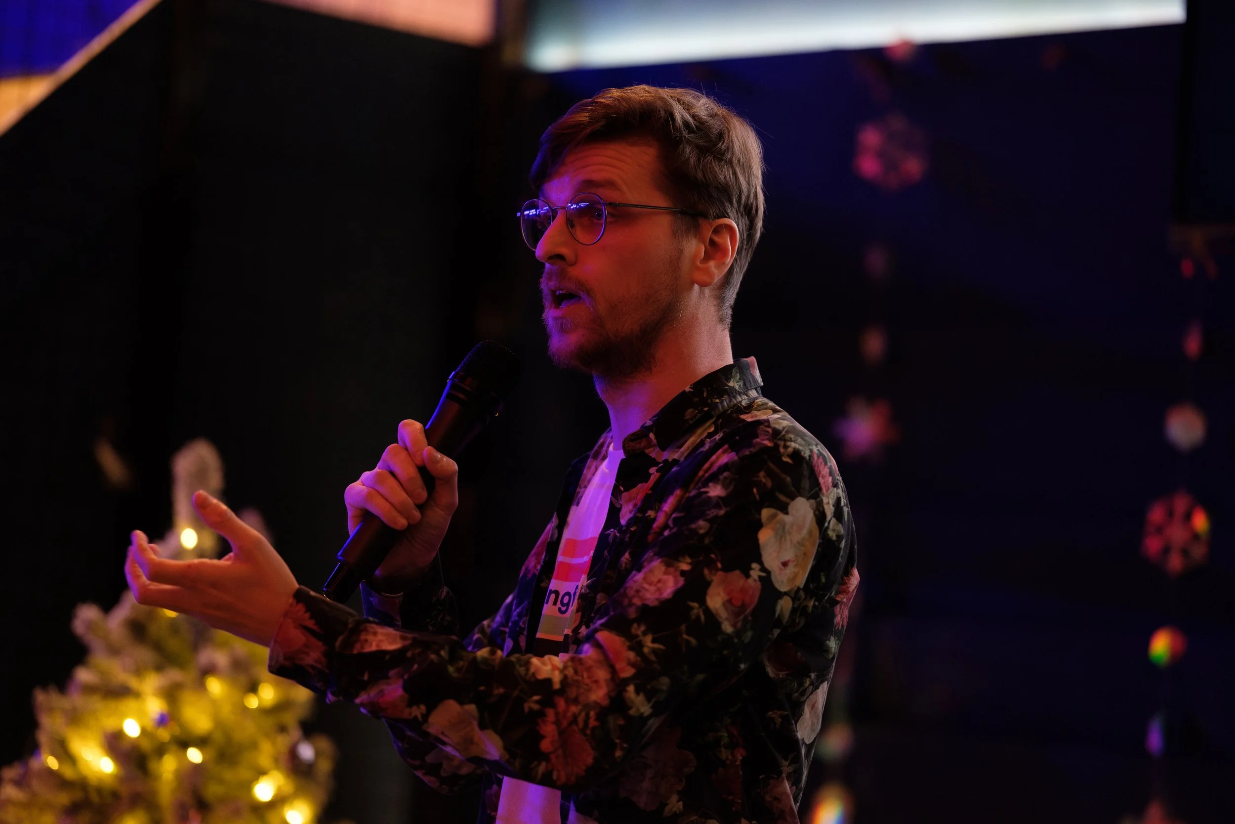 Person holding a microphone on stage with decorative lights in the background.