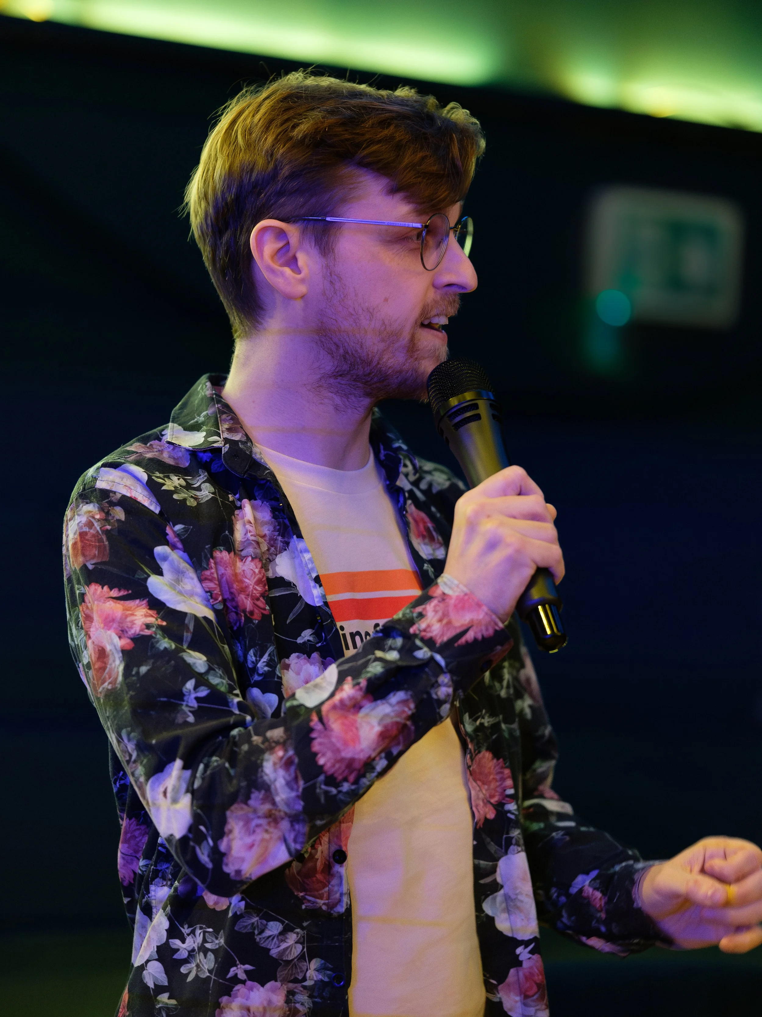 Person speaking into a microphone wearing glasses and a floral-patterned shirt.