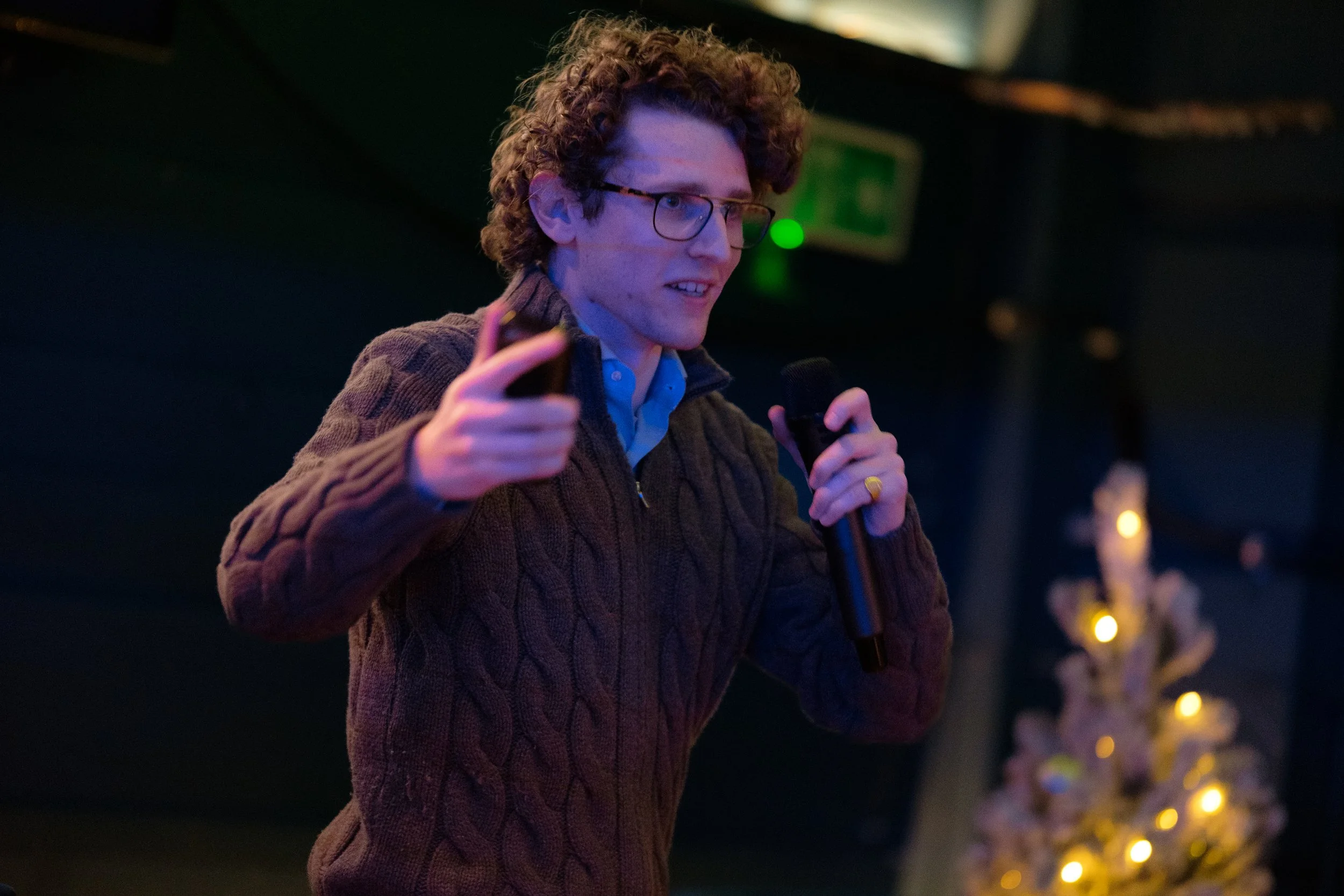 Person speaking with a microphone in one hand and gesturing with the other, wearing a sweater and glasses, with a Christmas tree decorated with lights in the background.