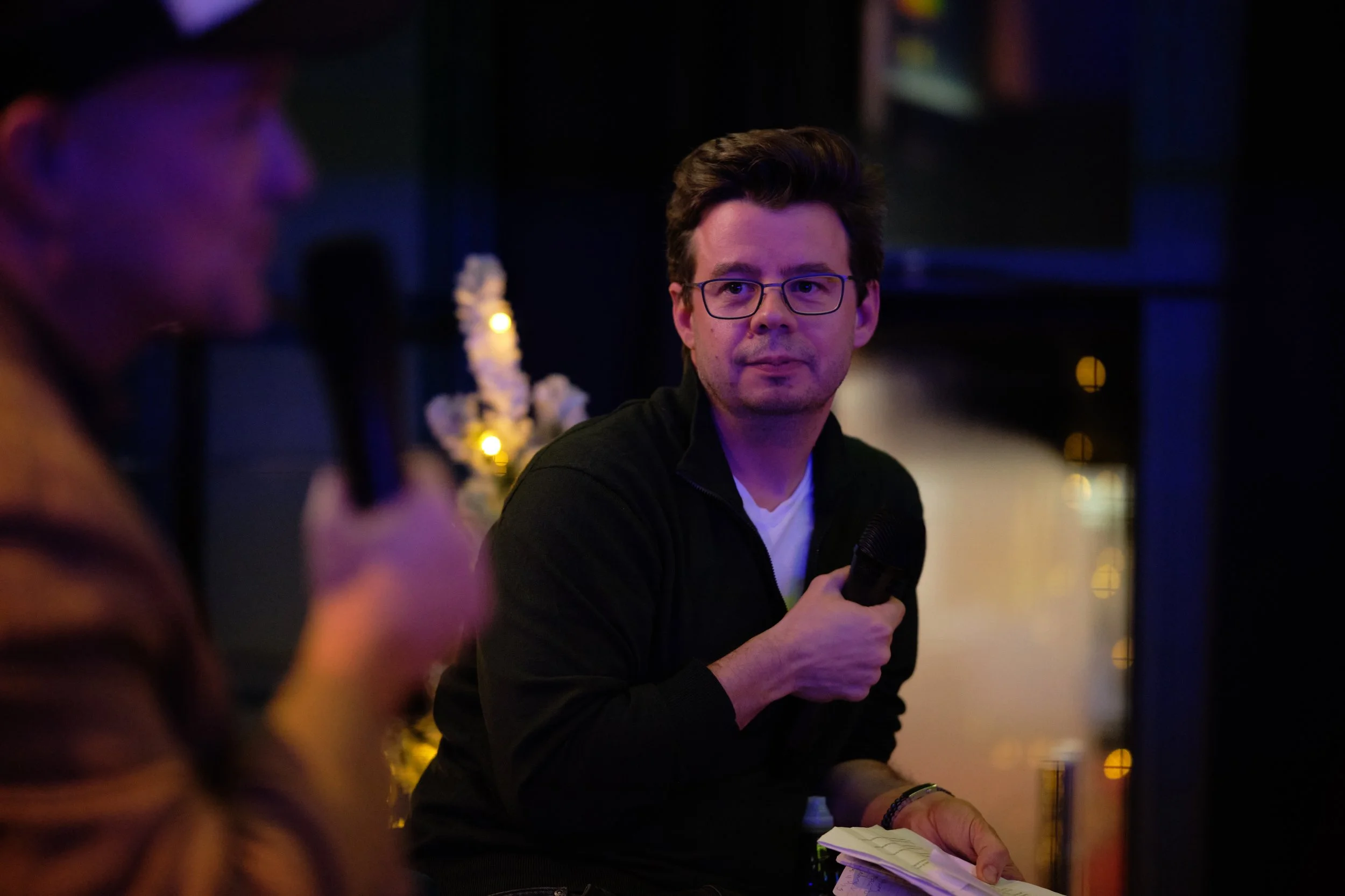 Two men holding microphones, engaged in a conversation indoors with a blurred background and dim lighting.