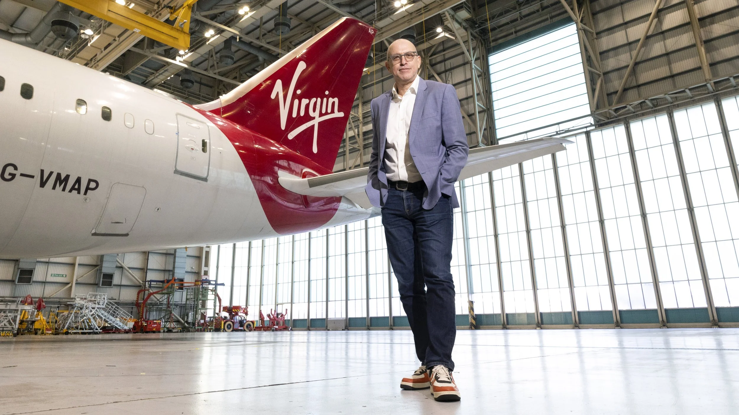 Shai Weiss outgoing CEO of Virgin Atlantic wears a light purple blazer, white shirt, and casual pants, standing inside an airplane hangar next to a Virgin Airlines airplane, with various equipment and tools visible in the background.
