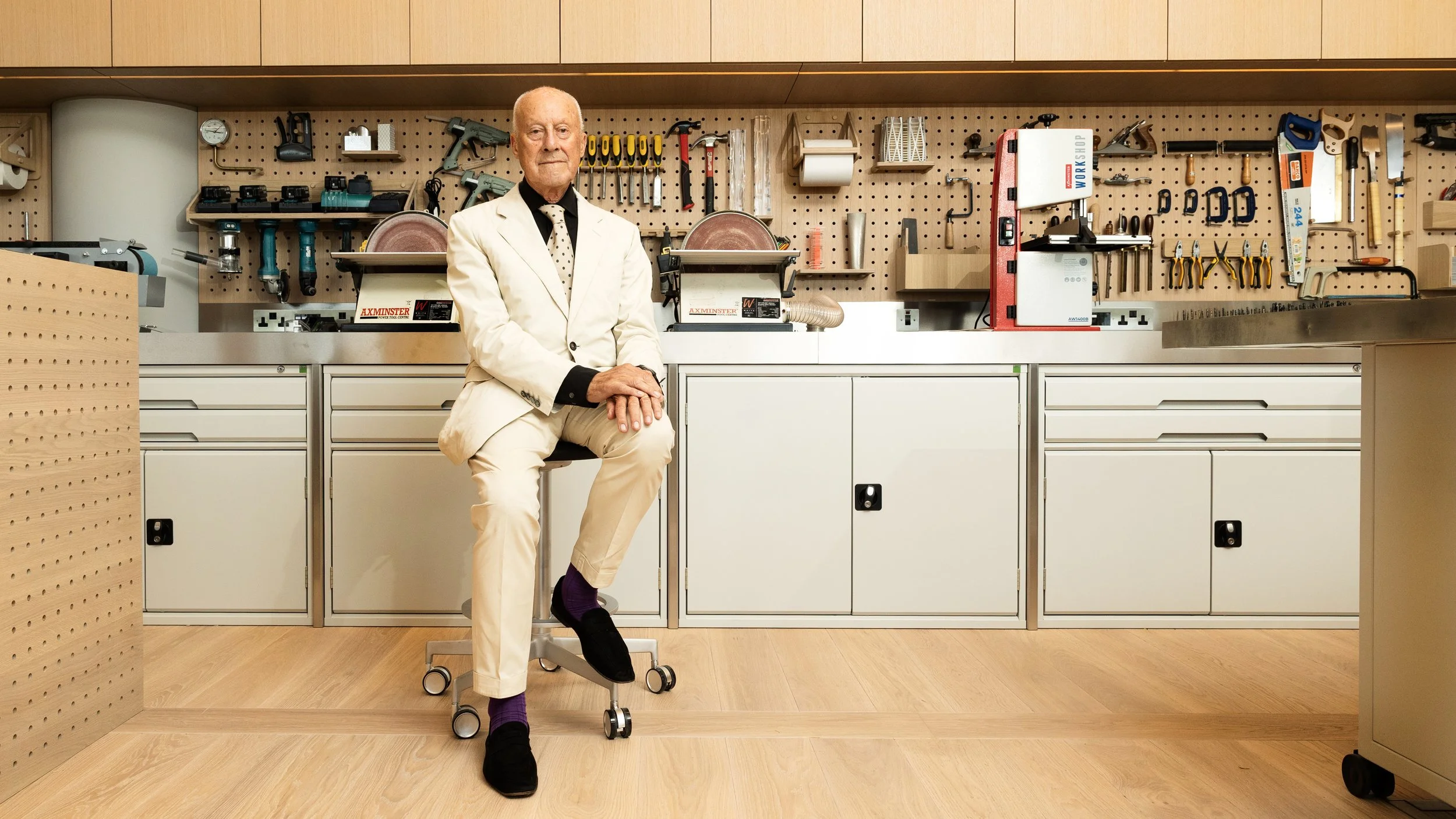 An older man in a white suit and black shoes sitting on a rolling stool in a workshop surrounded by tools and woodworking equipment.