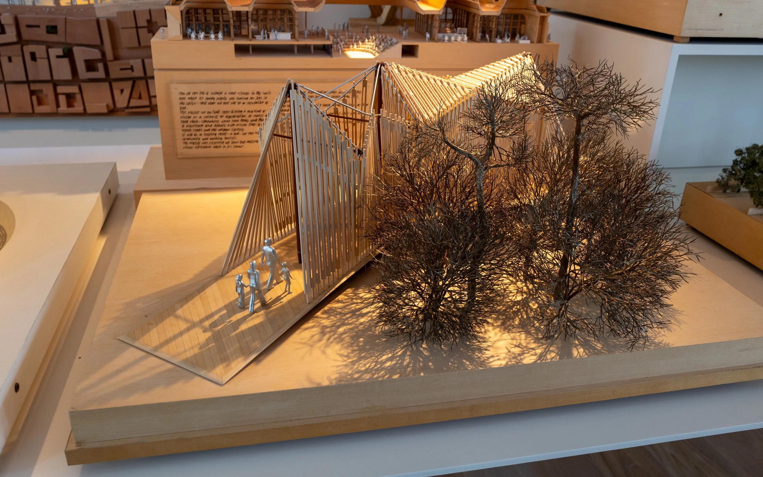 A detailed architectural model featuring a modern building with vertical wooden slats, surrounded by leafless trees and miniature human figures, set on a wooden base.