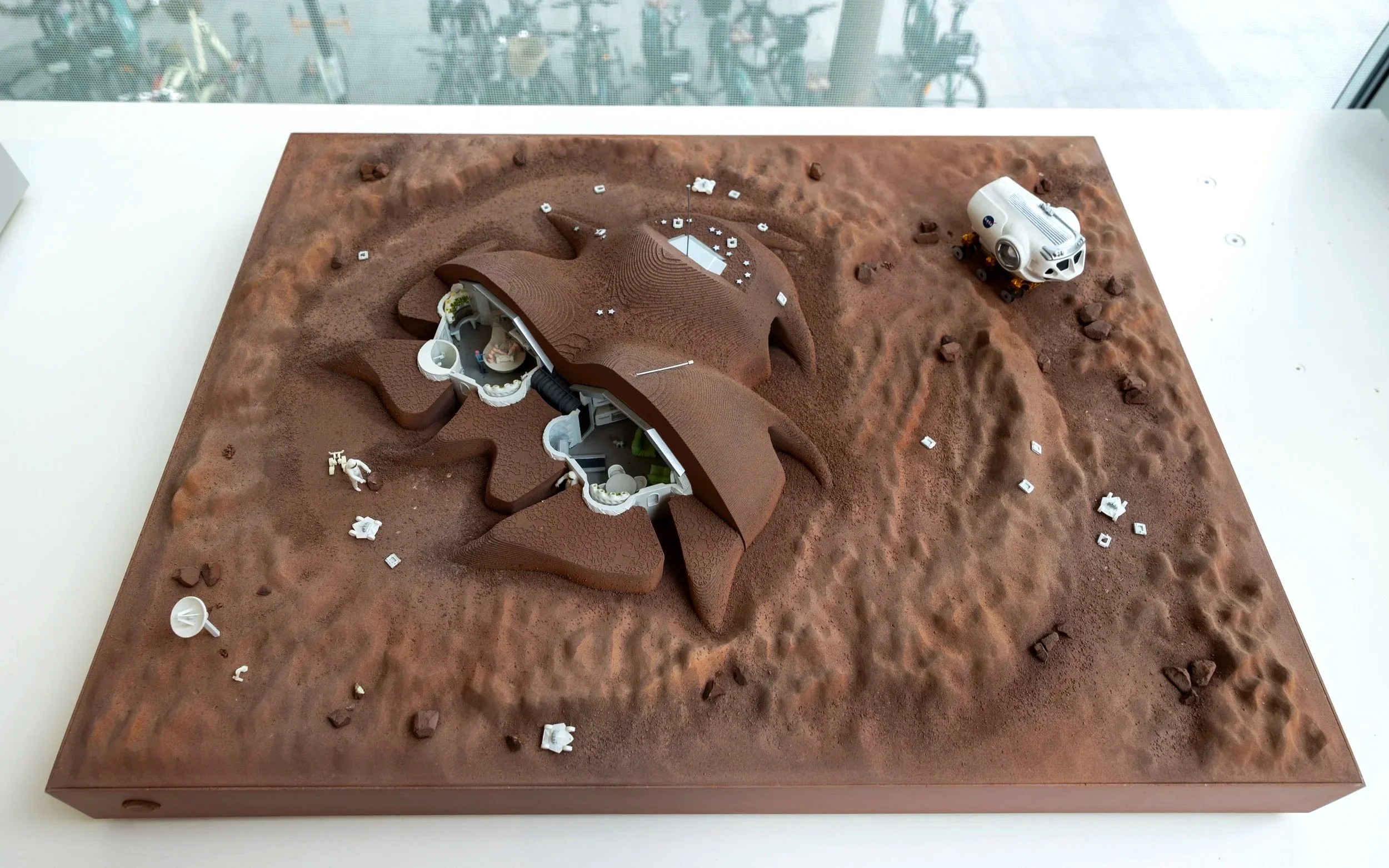 A detailed model of Mars terrain with mountains and craters, featuring small white structures, a white vehicle with NASA and space-related markings, and miniature astronauts and satellite dishes.