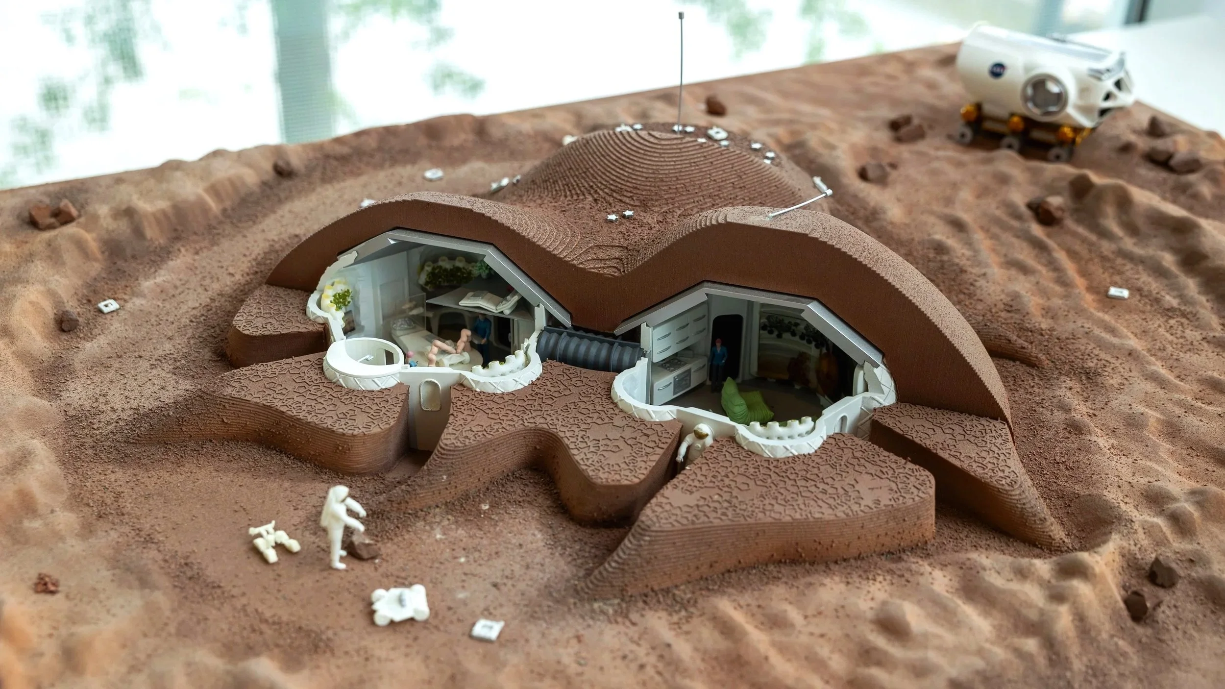 Cross-section of a Mars habitat model with detailed interior and roof terrain, set on a sandy surface with small scattered objects and a distant vehicle