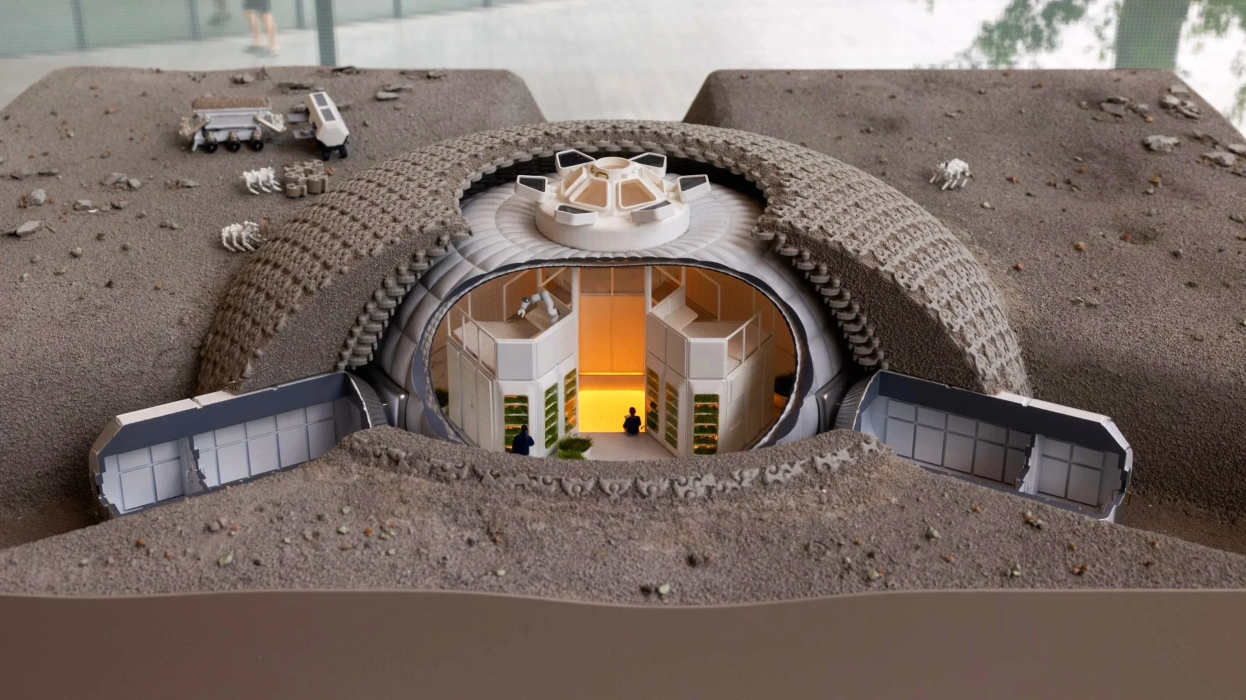 A detailed architectural model of a futuristic building with an underground entrance, surrounded by a textured terrain with small figures, vehicles, and scattered animals.