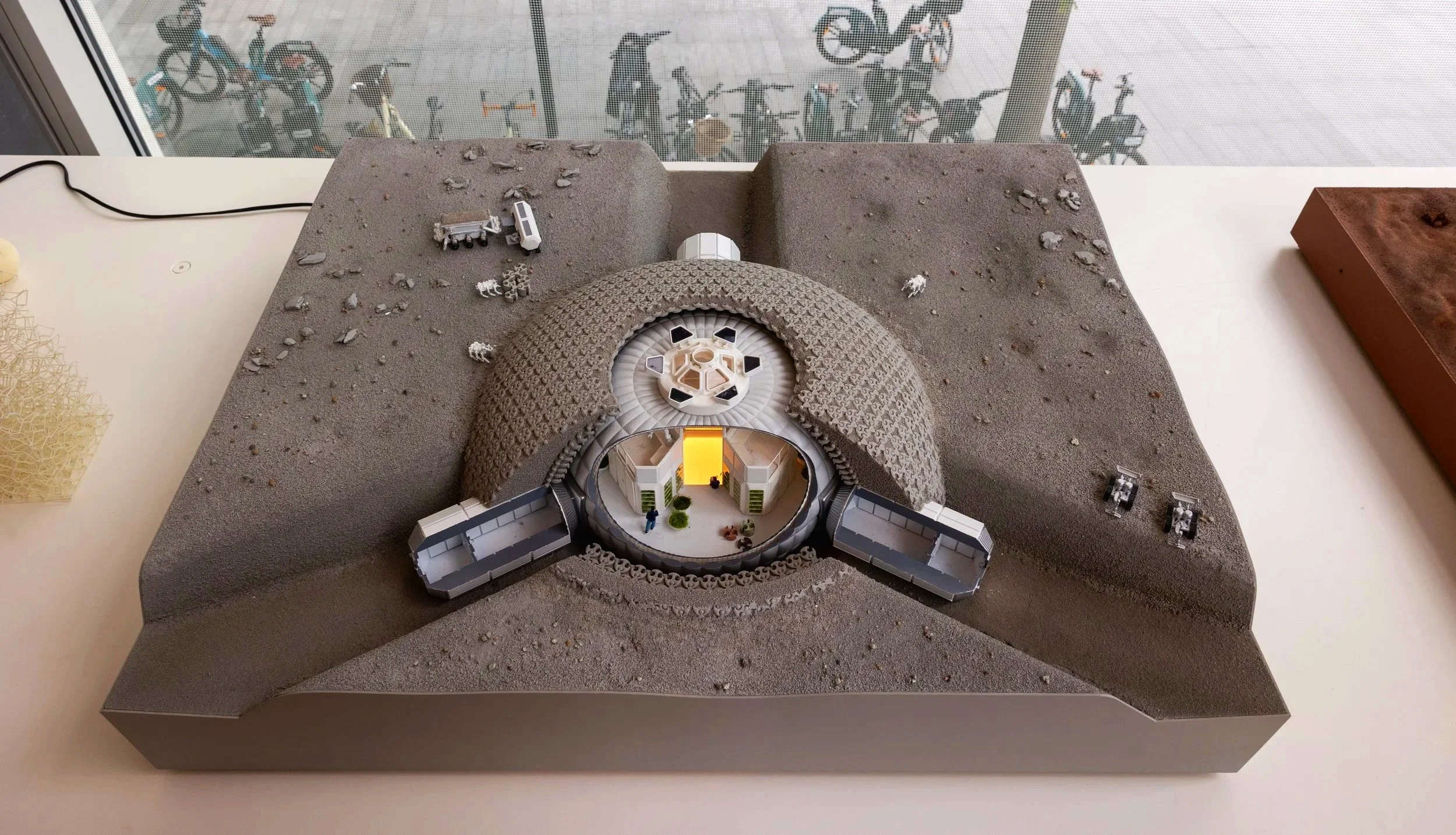 A detailed architectural model of a futuristic building with a domed entrance, surrounded by a textured landscape, on a white table. In the background, a window with a view of bicycles outside.