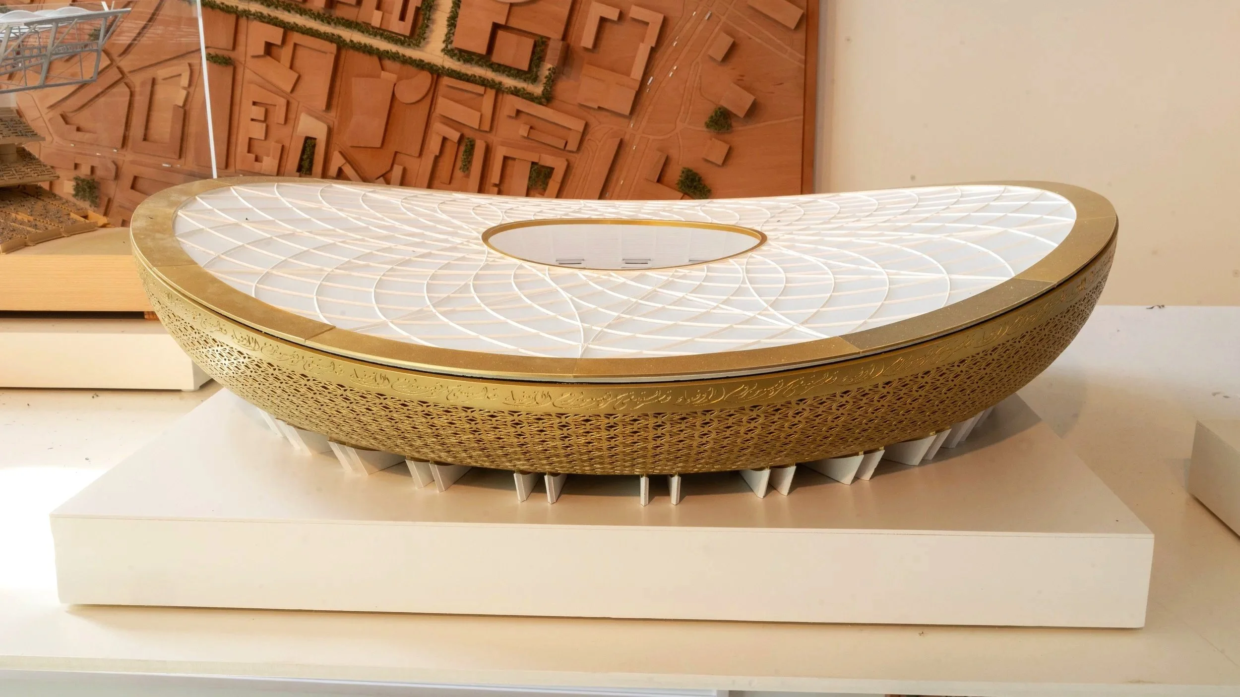 A detailed architectural model of a modern building with a gold and white exterior, featuring a unique curved design and decorative patterns on the sides, placed on a white platform.
