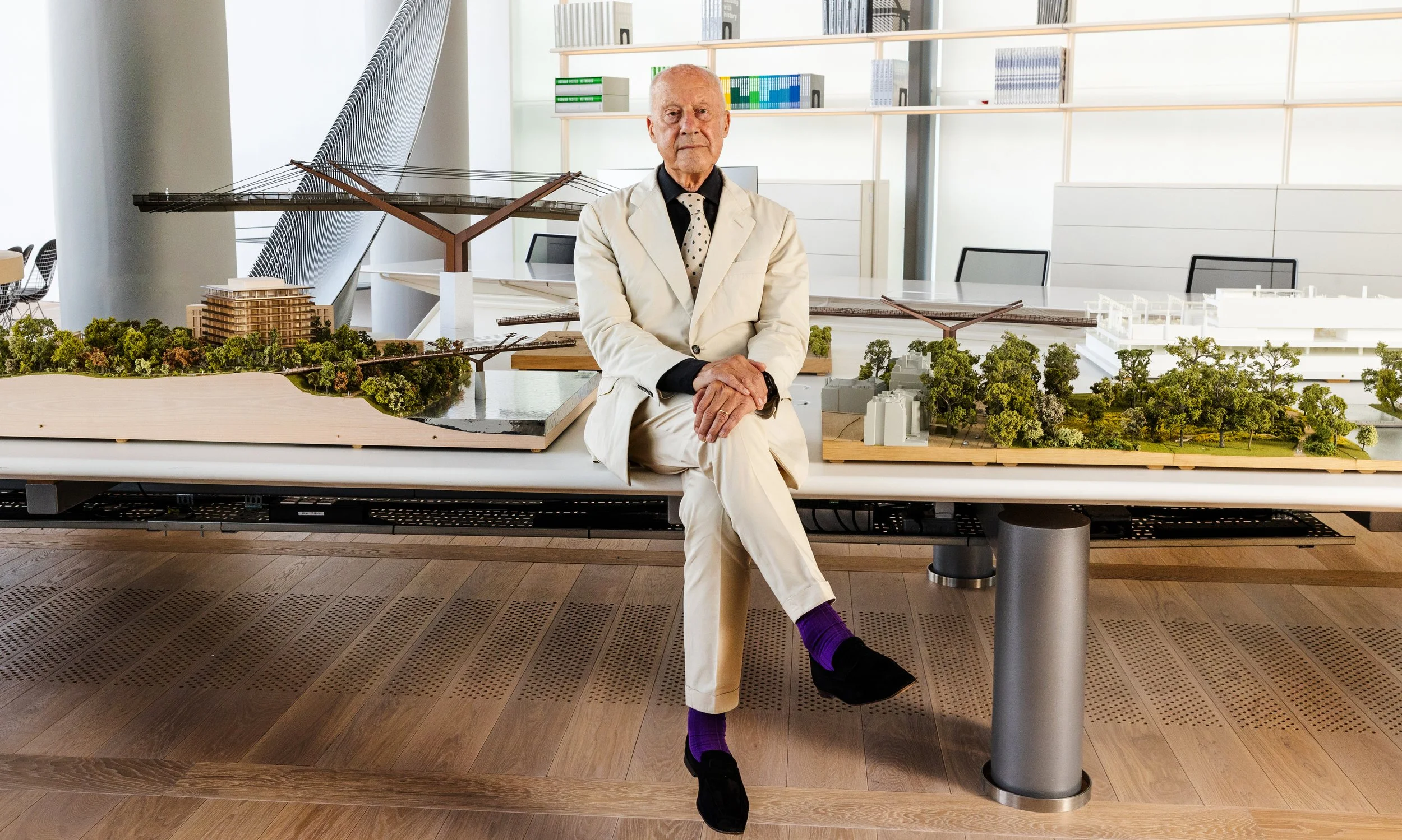 An elderly man in a white suit with a black shirt, polka dot tie, and purple socks, sitting on a table with architectural models and a miniature bridge, in a modern office or gallery space.