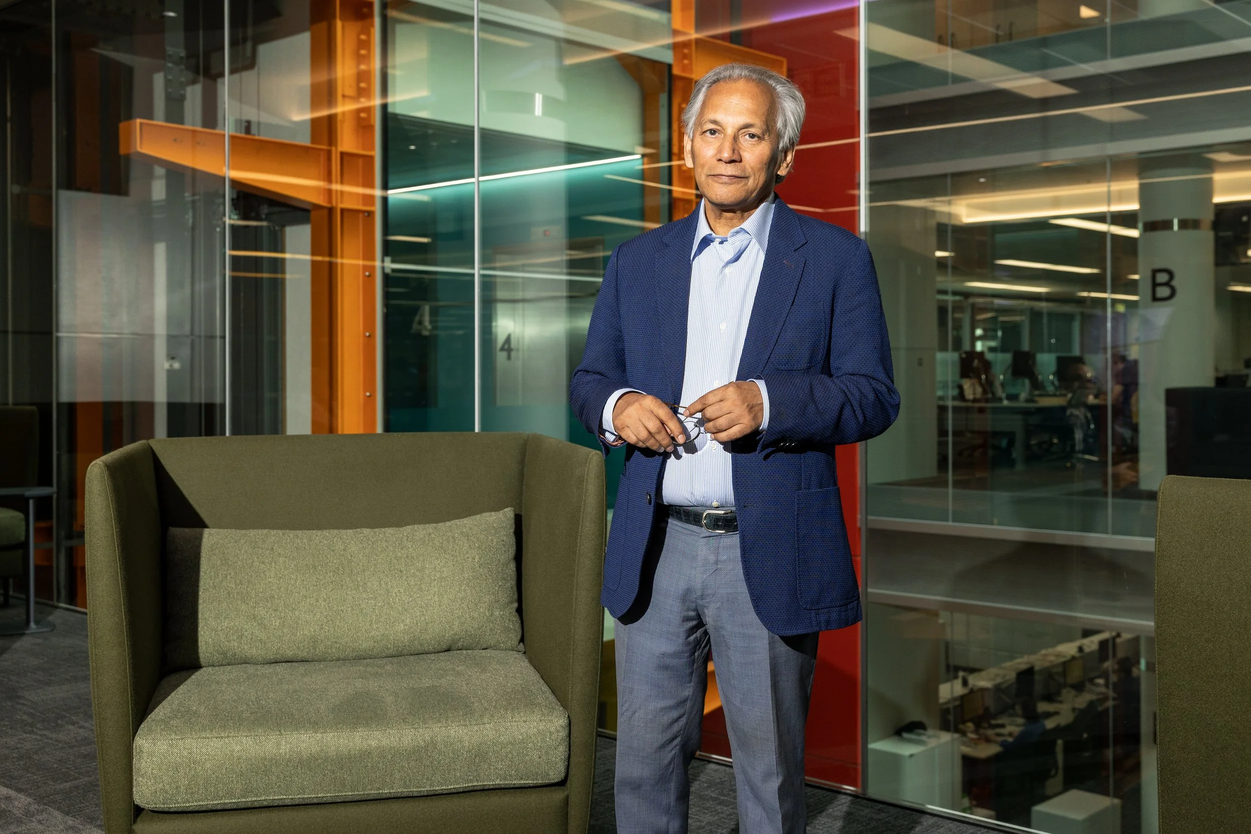 Dr Samir Shah CBE and Chairman of the BBC wears a blue blazer and gray pants, standing in an office lobby holding glasses.