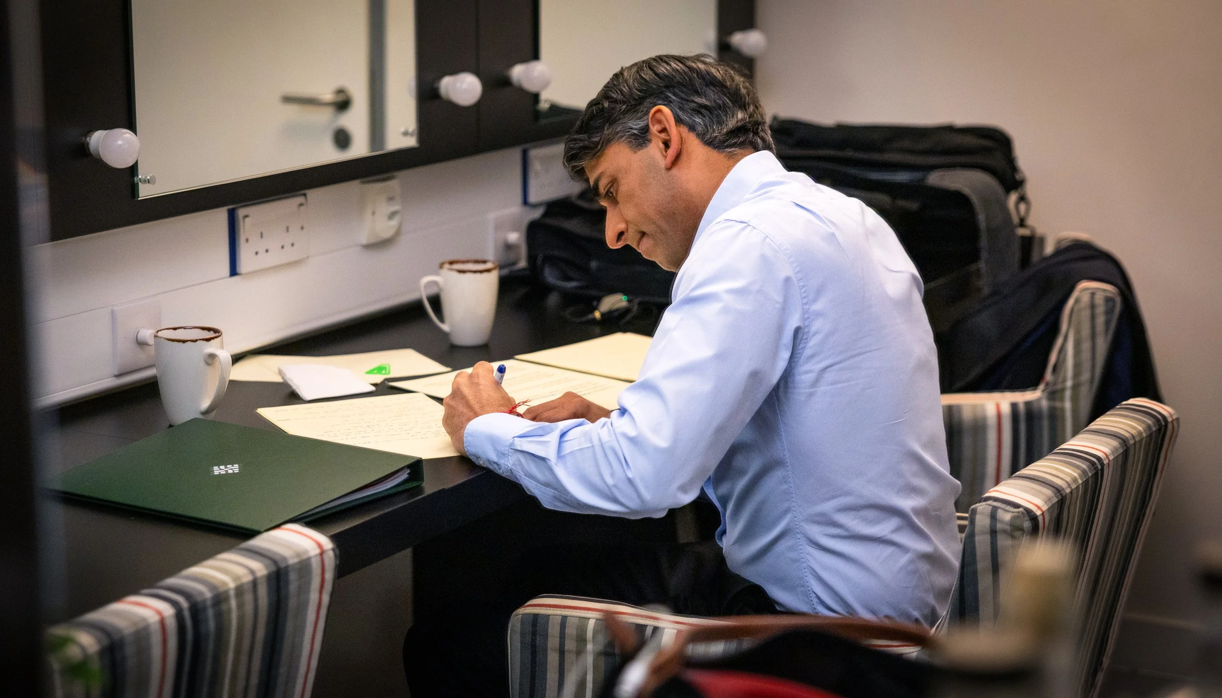 Ex-Prime minister Rishi Sunak sitting at a desk, writing on papers with a pen. There are two coffee mugs, a closed green binder, and a black backpack on the desk. The room has a mirror with several light bulbs and electrical outlets on the wall.