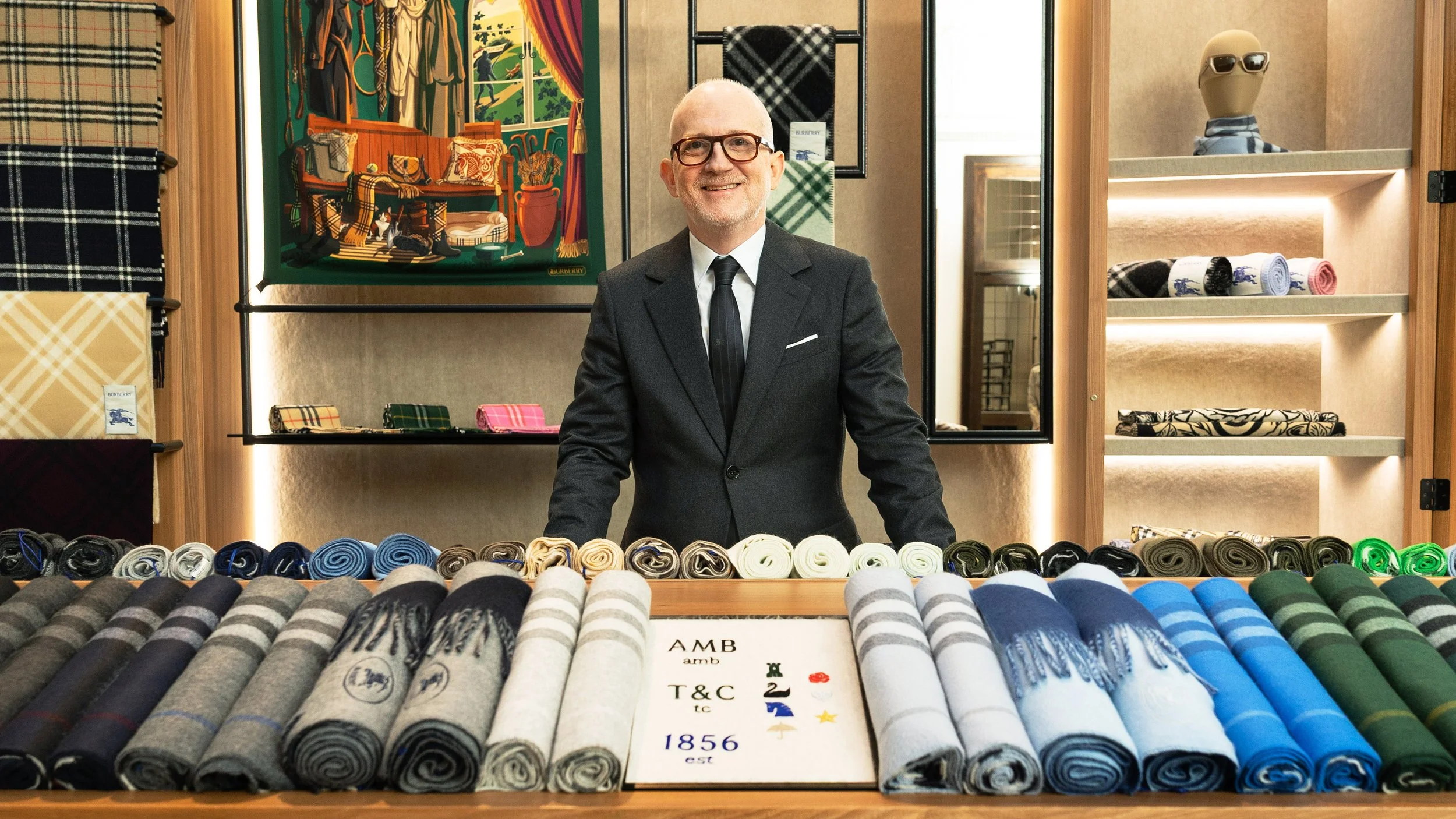 Burberry CEO Joshua Shulman wears a
 black suit and tie standing behind a display of rolled fabric in various colors and patterns, with shelves of fabric and a framed artwork in the background.