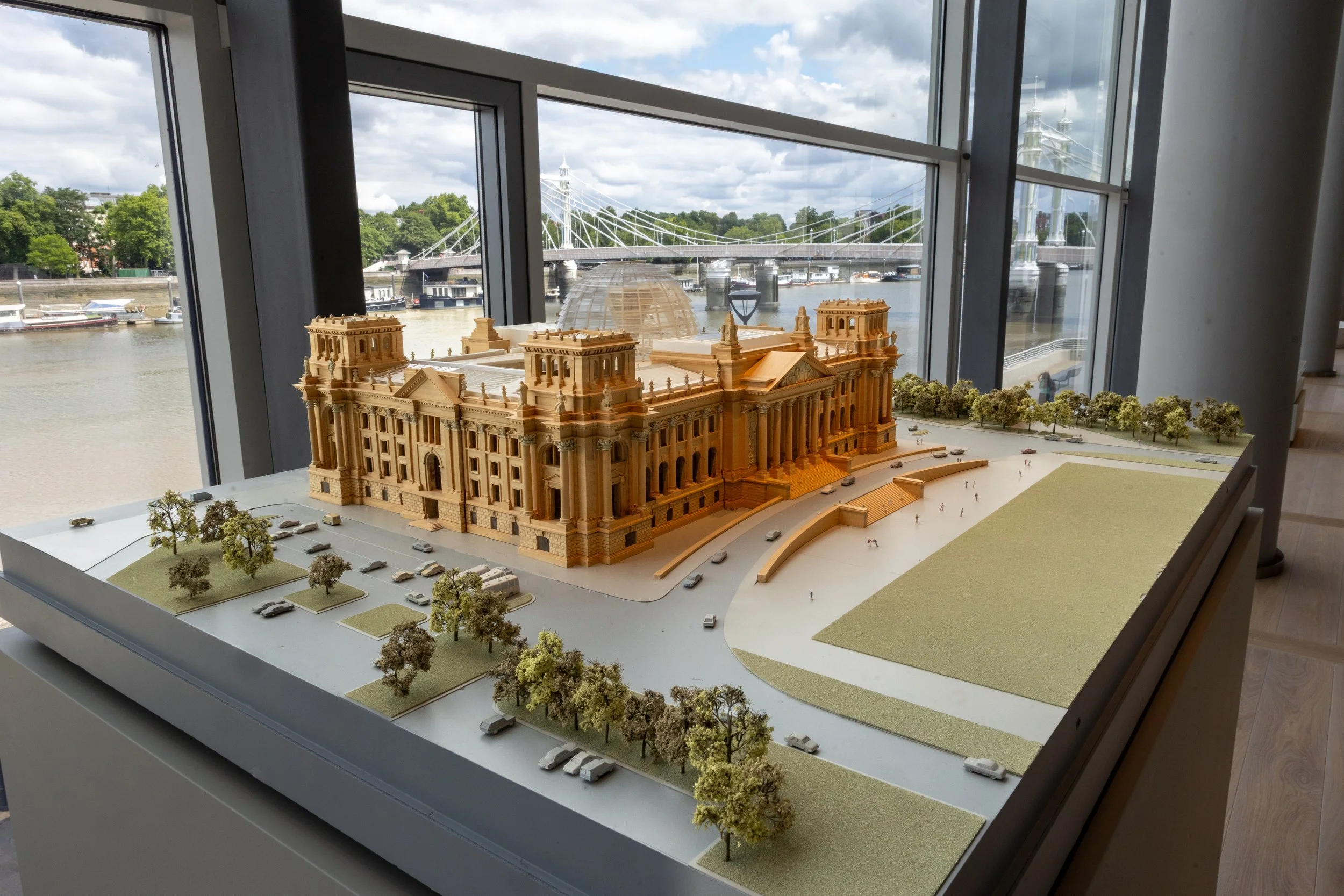 A detailed architectural scale model of a historic building with trees and parking areas in the foreground, set inside a building with large windows overlooking a river and a bridge in the background.