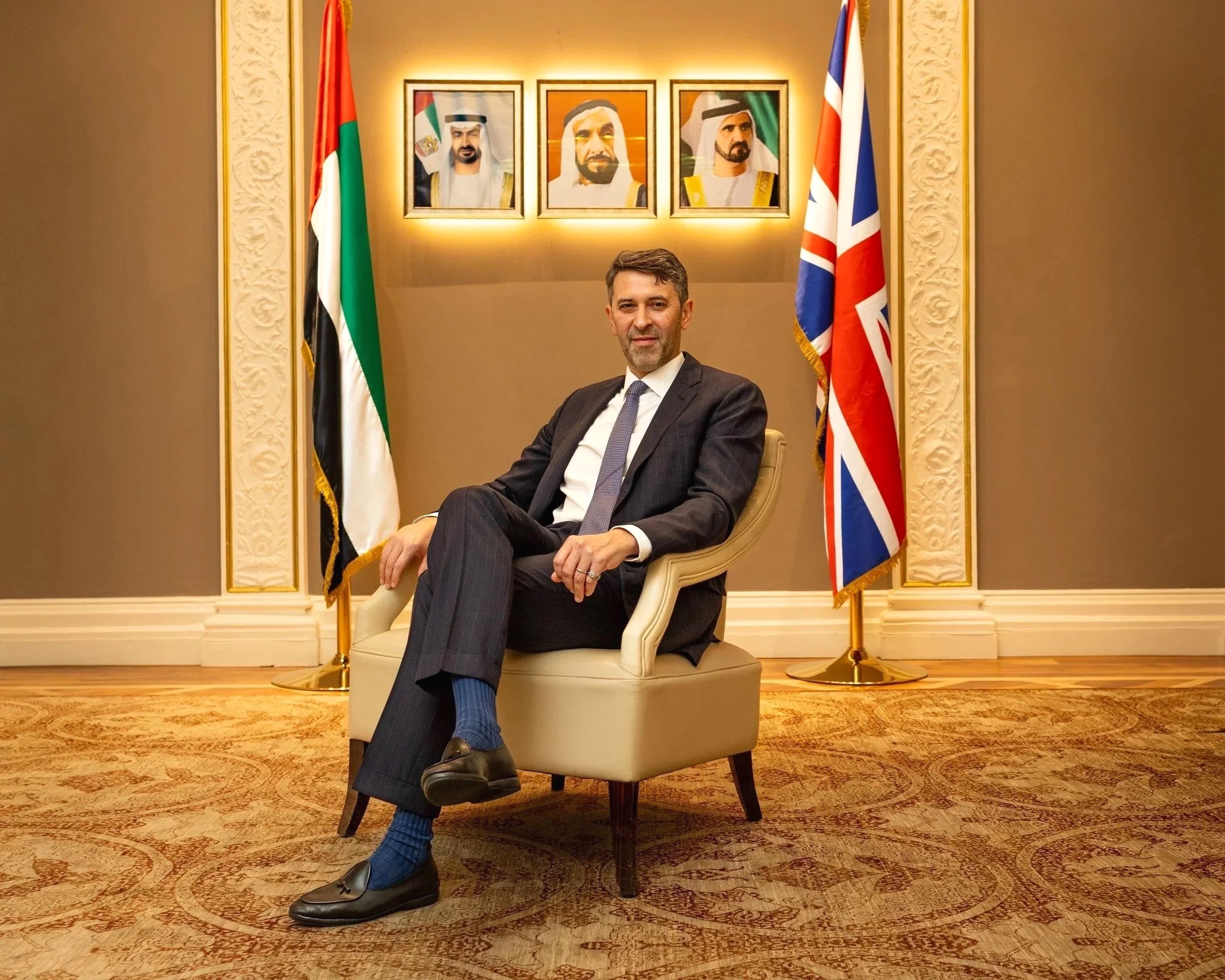 A man in a suit sitting comfortably in an armchair in a formal room decorated with portraits of Middle Eastern leaders and flags of the United Arab Emirates and the United Kingdom.