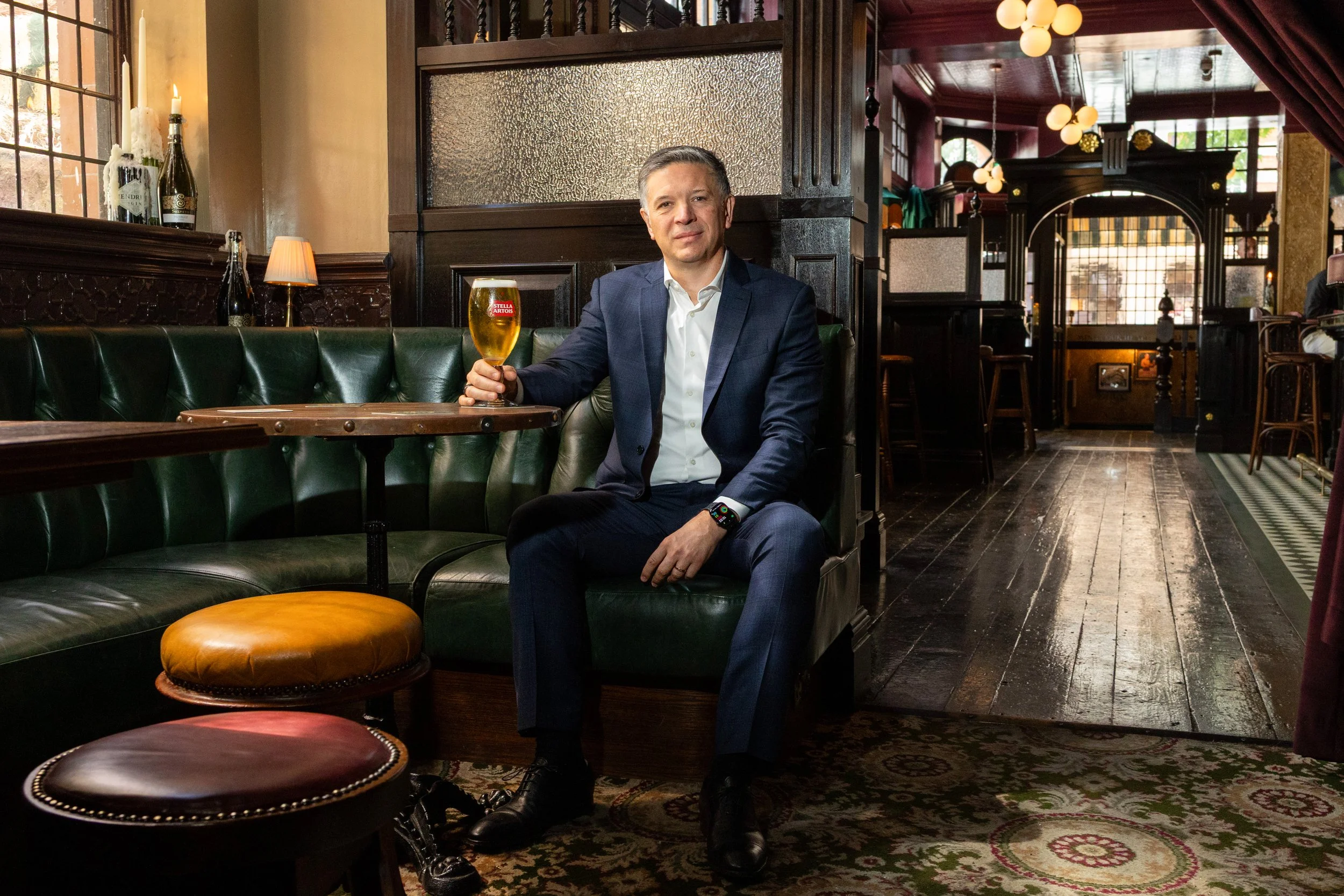 CEO of AB InBev Michel Doukeris sits on a green leather booth in a vintage-style restaurant, holding a glass of Stella Artois beer. The interior features dark wood furniture, dim lighting, decorative bulbs, and a patterned carpet.