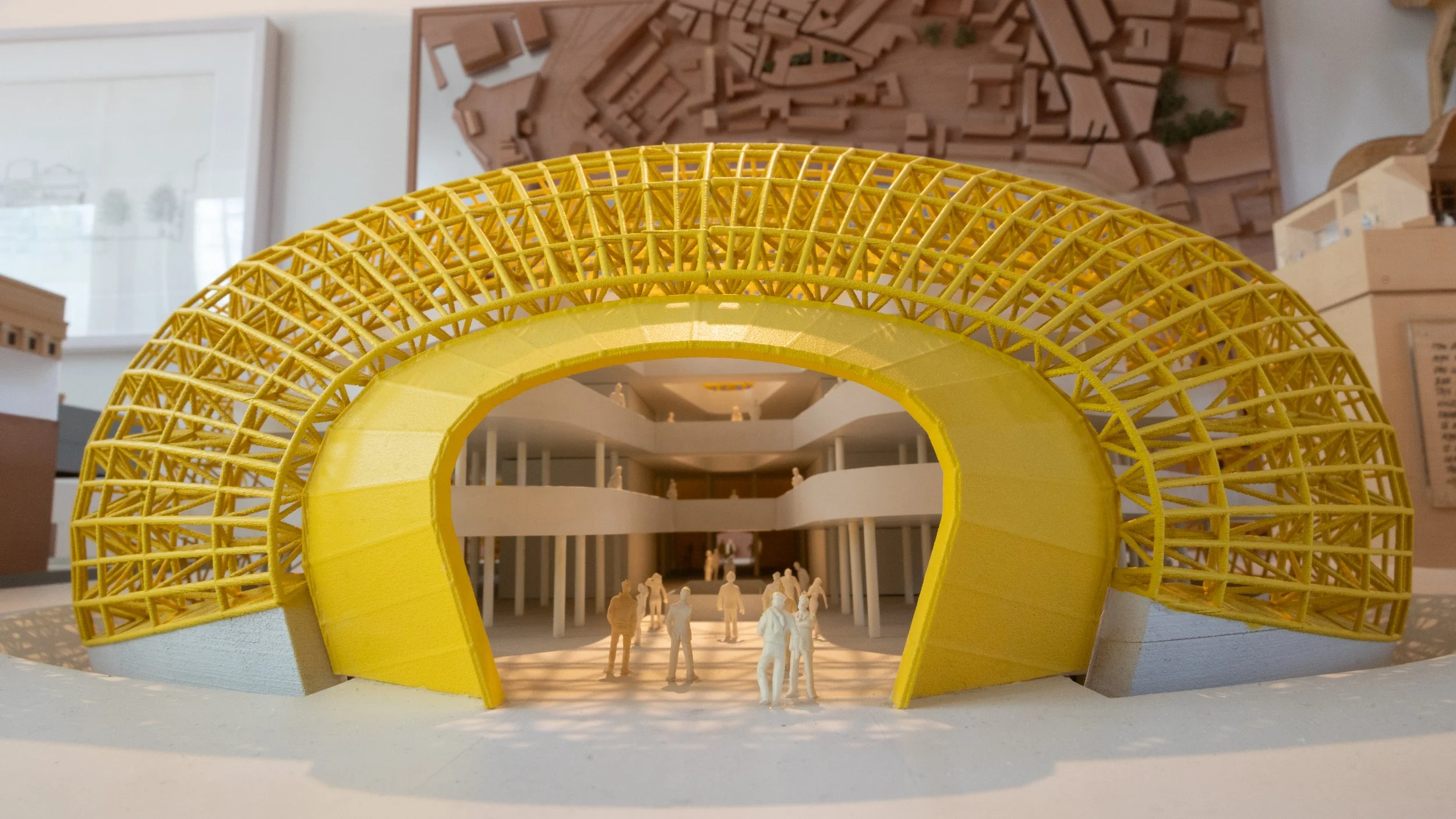 A yellow architectural model of a circular building with a lattice roof structure on display, with miniature figures inside and a background of drawings and plans.