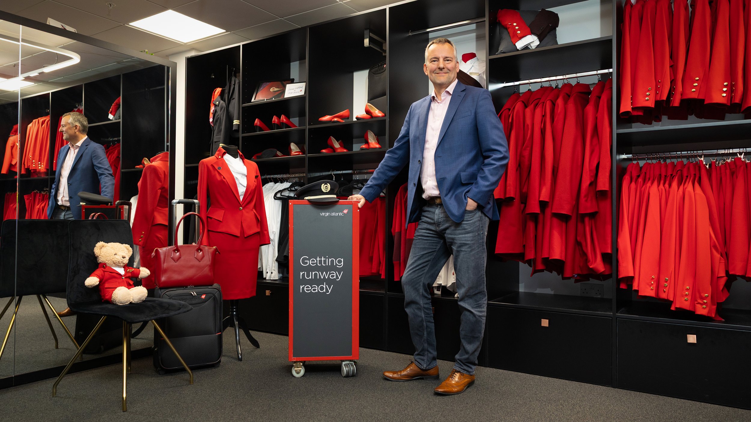 CEO of Virgin Atlantic Corneel Koster wears a blue blazer and jeans standing in front of a display of red uniforms and accessories, with a teddy bear and bags in the foreground. The display includes a sign that says 'Getting runway ready'.