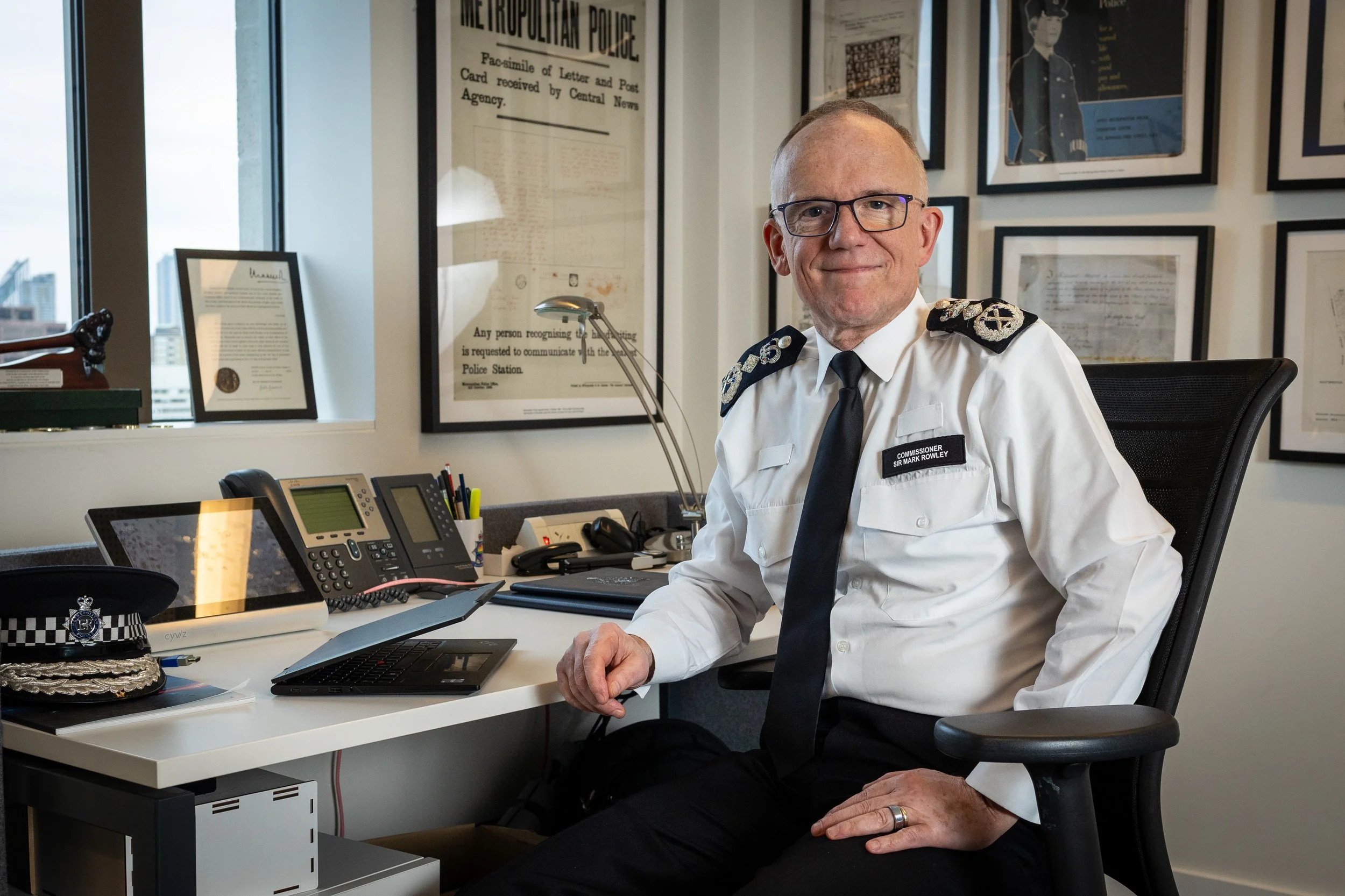 MET Commissionofficer wearing glasses, a white uniform with a badge that reads 'Commissioner Sir Mark Rowley sitting at his desk in an office decorated with framed pictures and documents. There is a police hat and various office supplies on the desk.