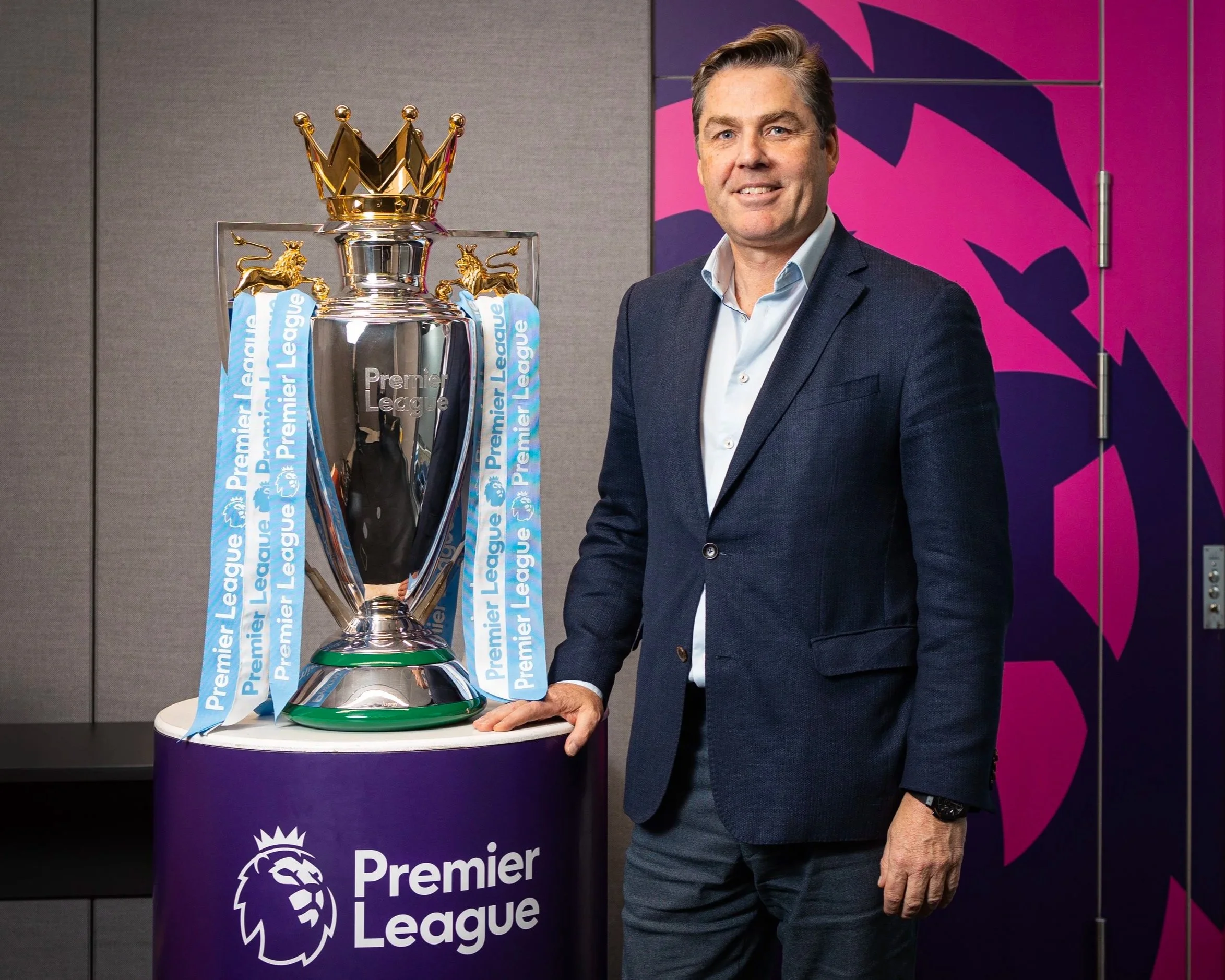 Richard Master CEO of The Premier League wears a suit standing next to a trophy with a gold crown on top, lions on the sides, and ribbons that say 'Premier League,' with a purple banner in the background.