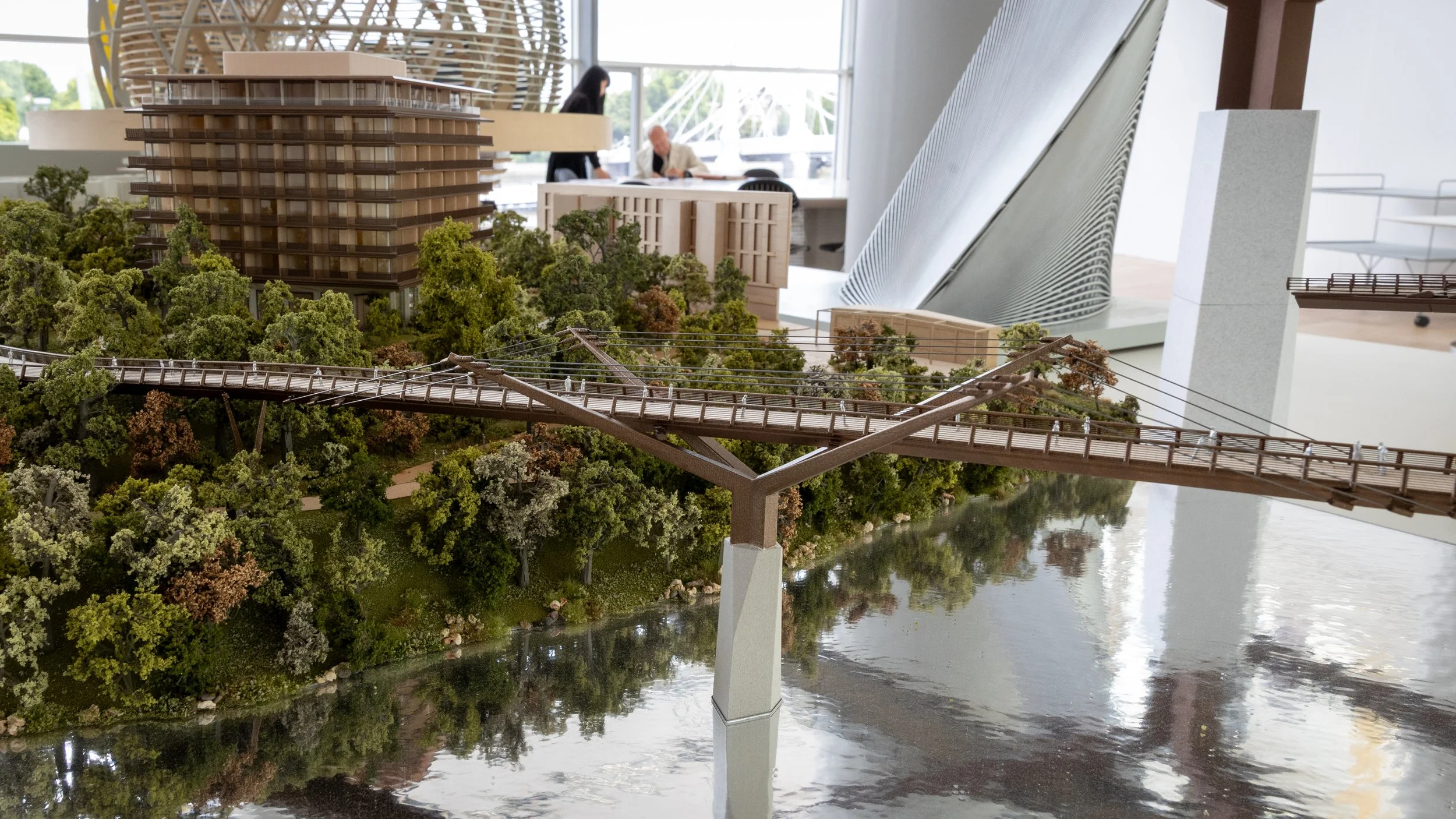 Model of an urban development project showing a bridge over a river, surrounded by trees, with modern buildings and an indoor setting in the background.