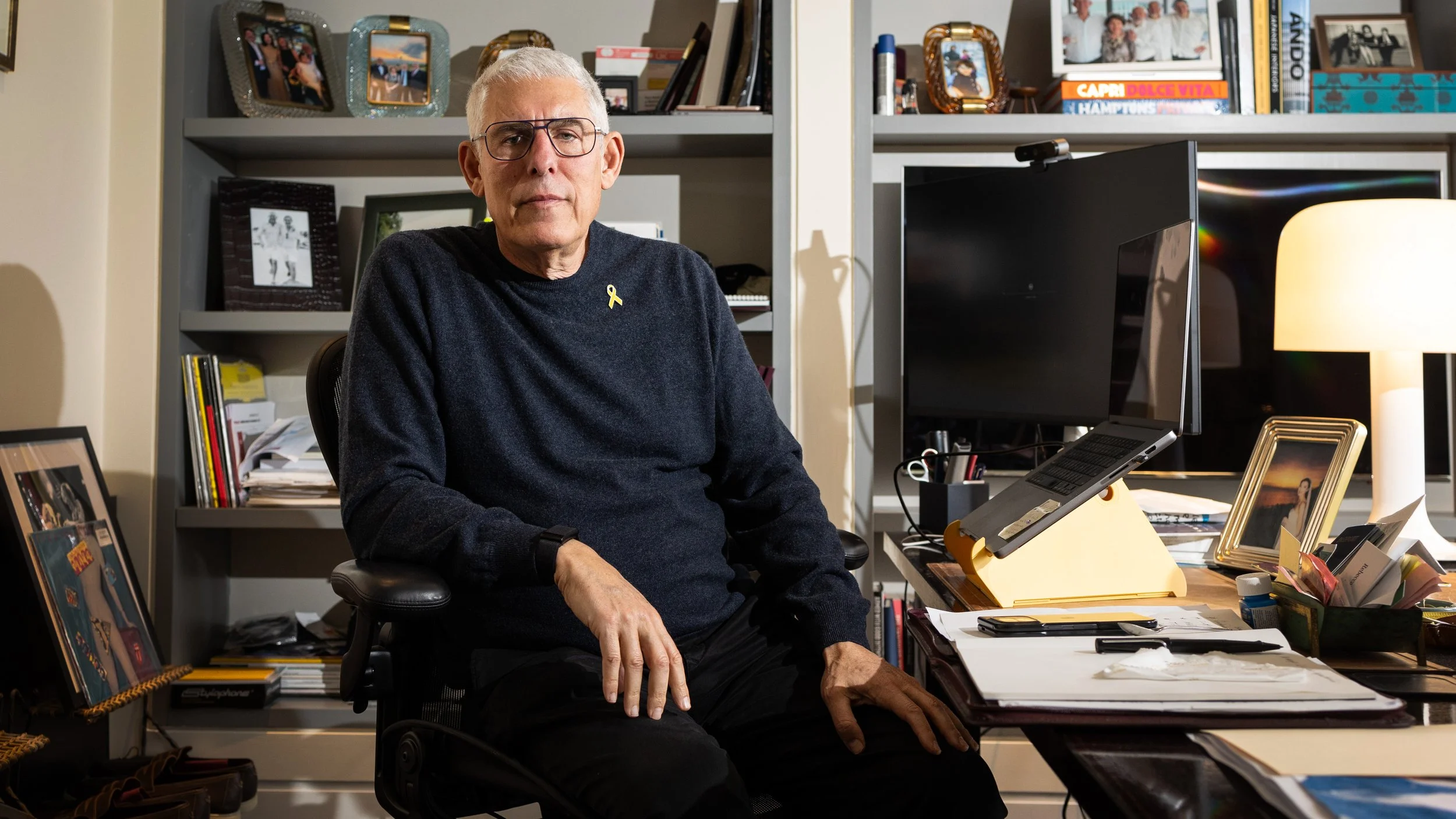 An older man with gray hair and glasses sitting in a black office chair at a cluttered desk, surrounded by photo frames, books, and a computer.